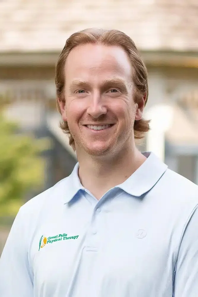 Smiling man wearing a light blue polo shirt with Great Falls Physical Therapy logo, standing outdoors with a blurred background of trees and a porch.