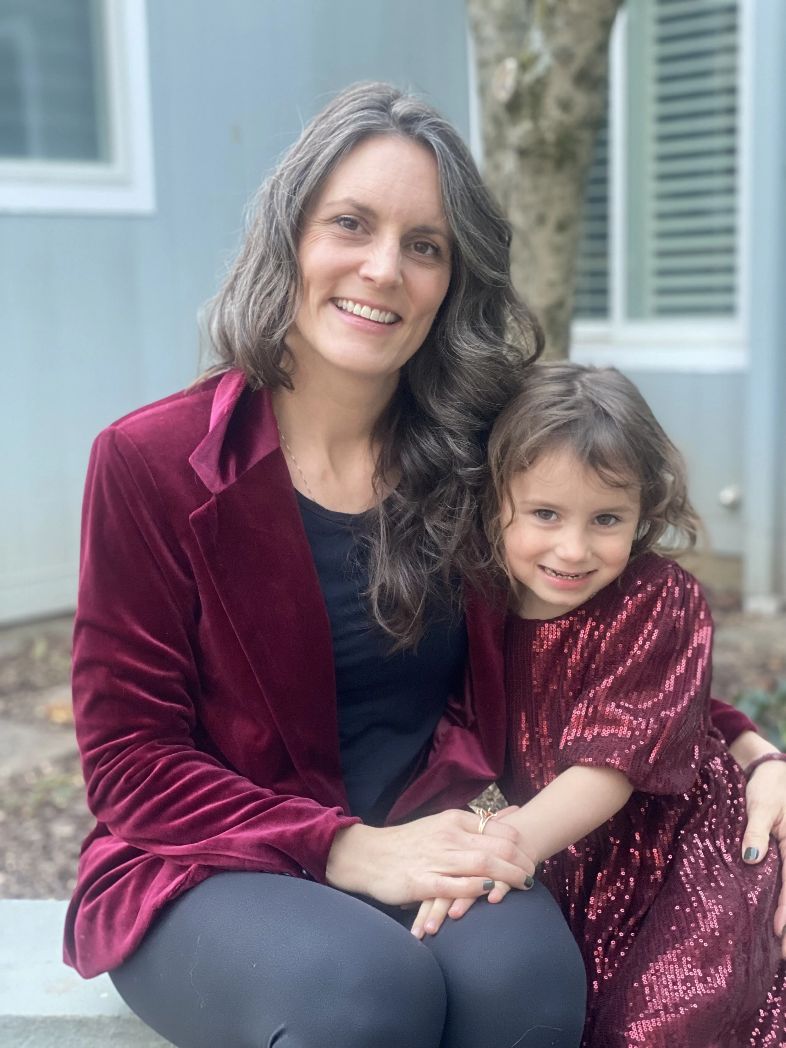 A woman and a young girl sitting outdoors, smiling at the camera. The woman has long, wavy gray hair and is wearing a red velvet jacket. The girl has shoulder-length curly brown hair and is dressed in a red, sequined dress.