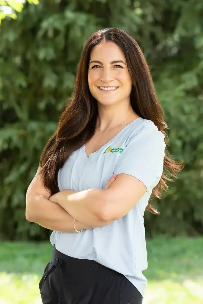 A woman with long brown hair smiling outdoors, wearing a light blue medical scrub top and black pants, with arms crossed, in front of green foliage.