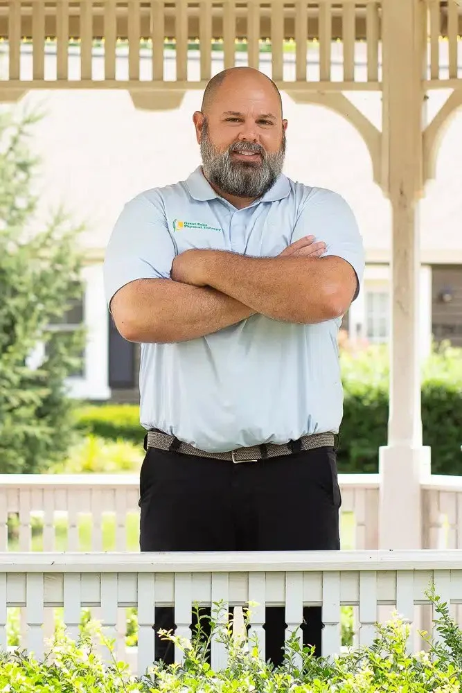A man with a beard, wearing a light blue collared shirt with a logo on it, standing outdoors with arms crossed.