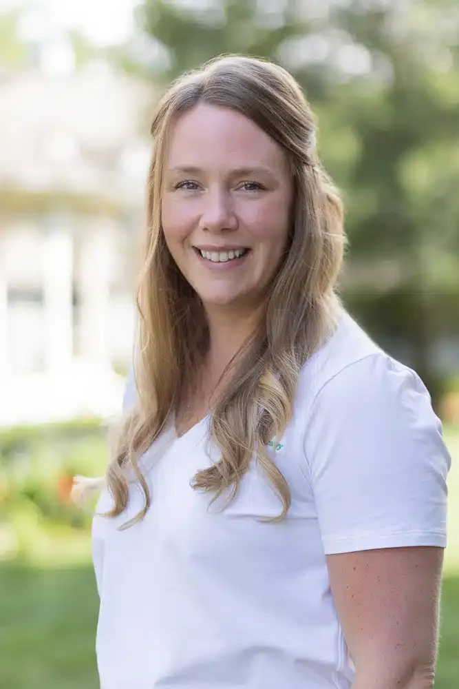A young woman with long, wavy blonde hair smiling outdoors in a white shirt.