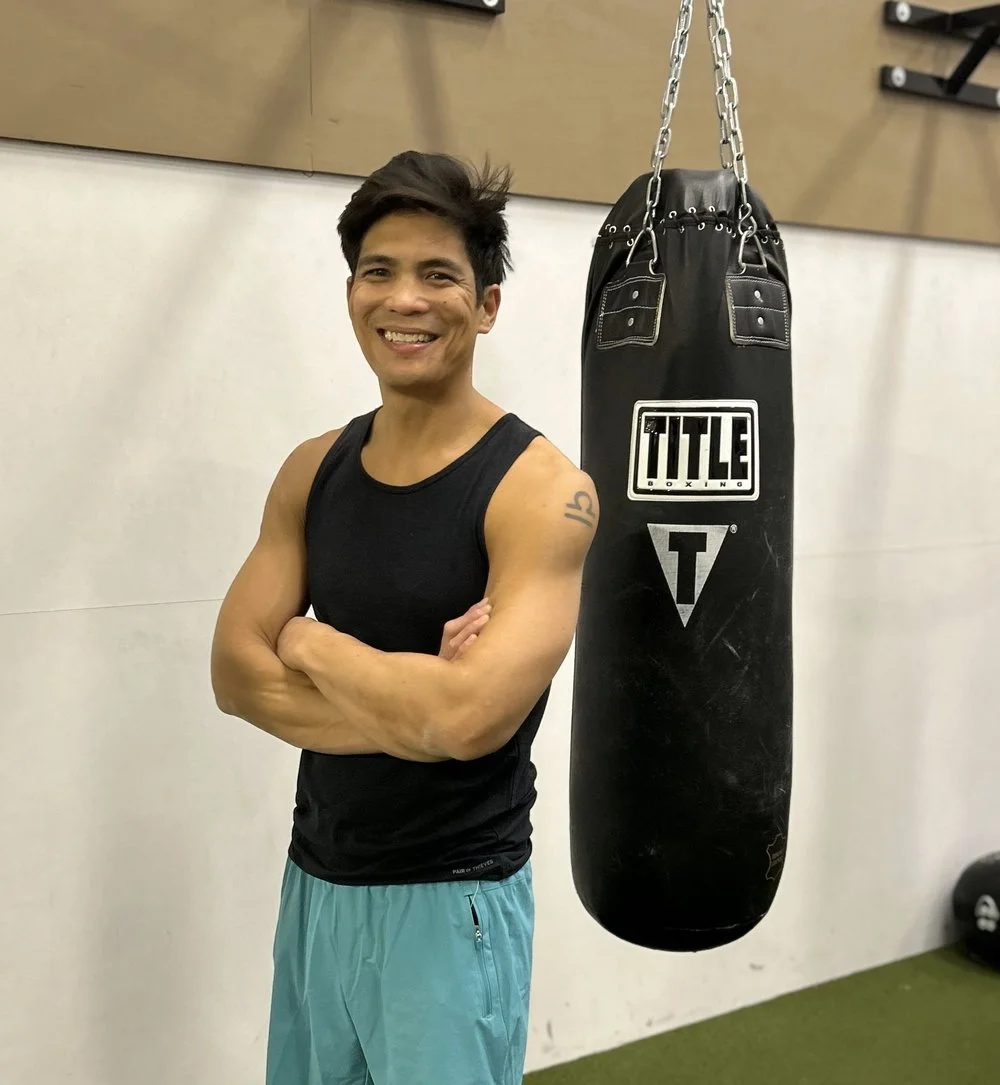A man in a black tank top and blue shorts smiling with arms crossed stands next to a hanging black heavy bag in a gym.