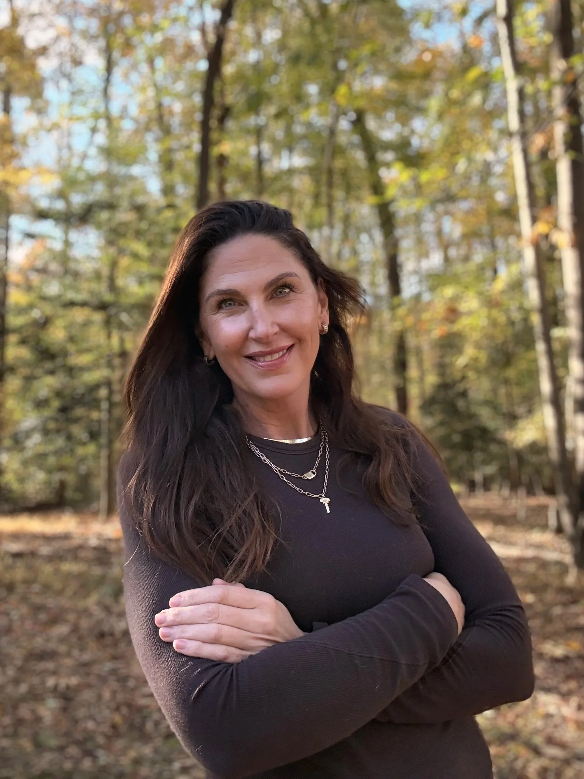 A woman with long dark hair standing outdoors in a forest during fall, smiling with arms crossed, wearing a black long-sleeve top and layered necklaces.