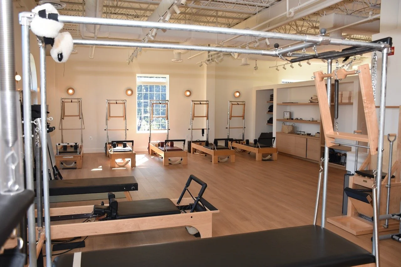 Pilates studio with wooden reformer machines arranged in rows, large windows allowing natural light, minimalist decor, and shelves with accessories.