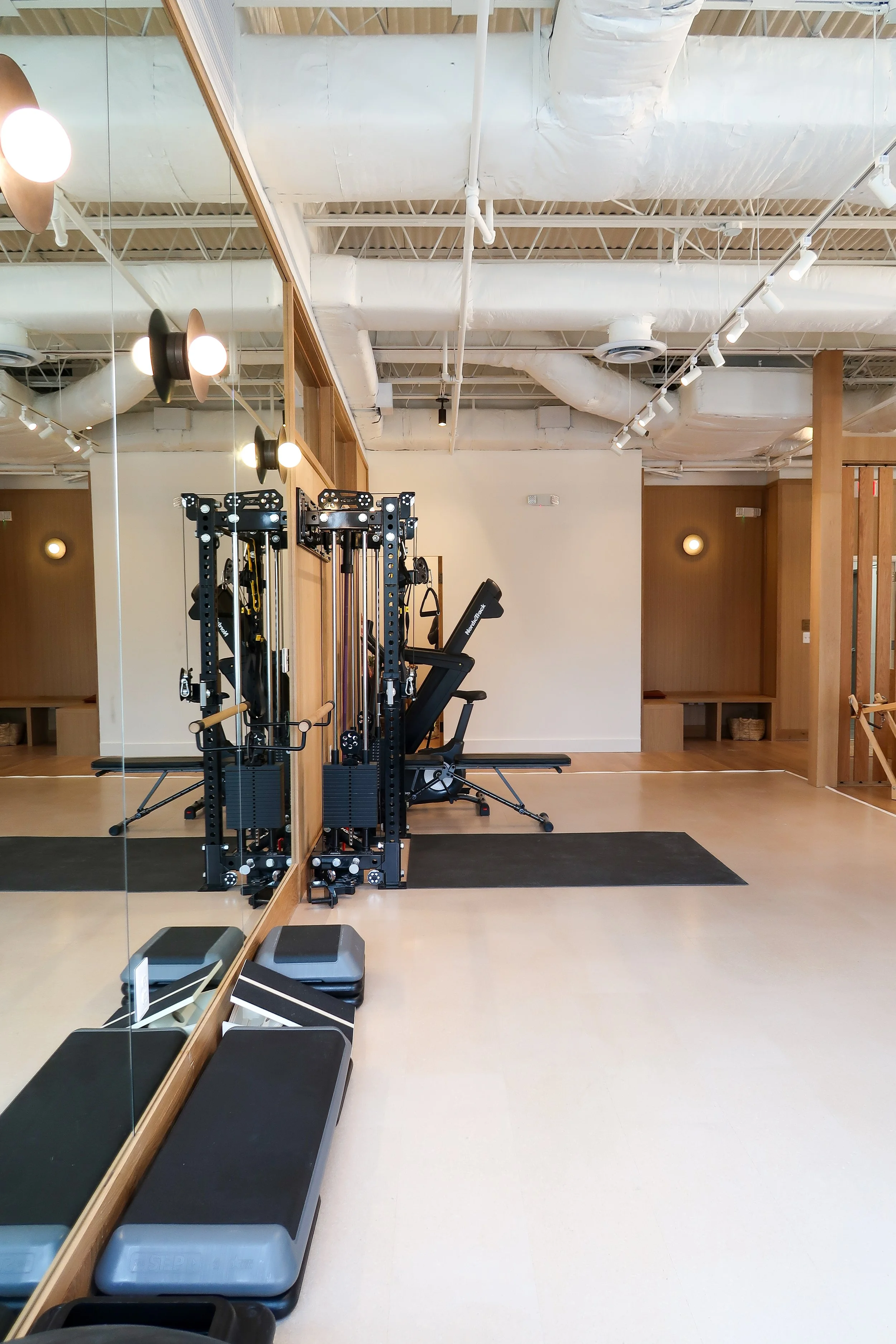Empty gym room with workout equipment, exercise benches, large mirror, black mats, and modern lighting fixtures.