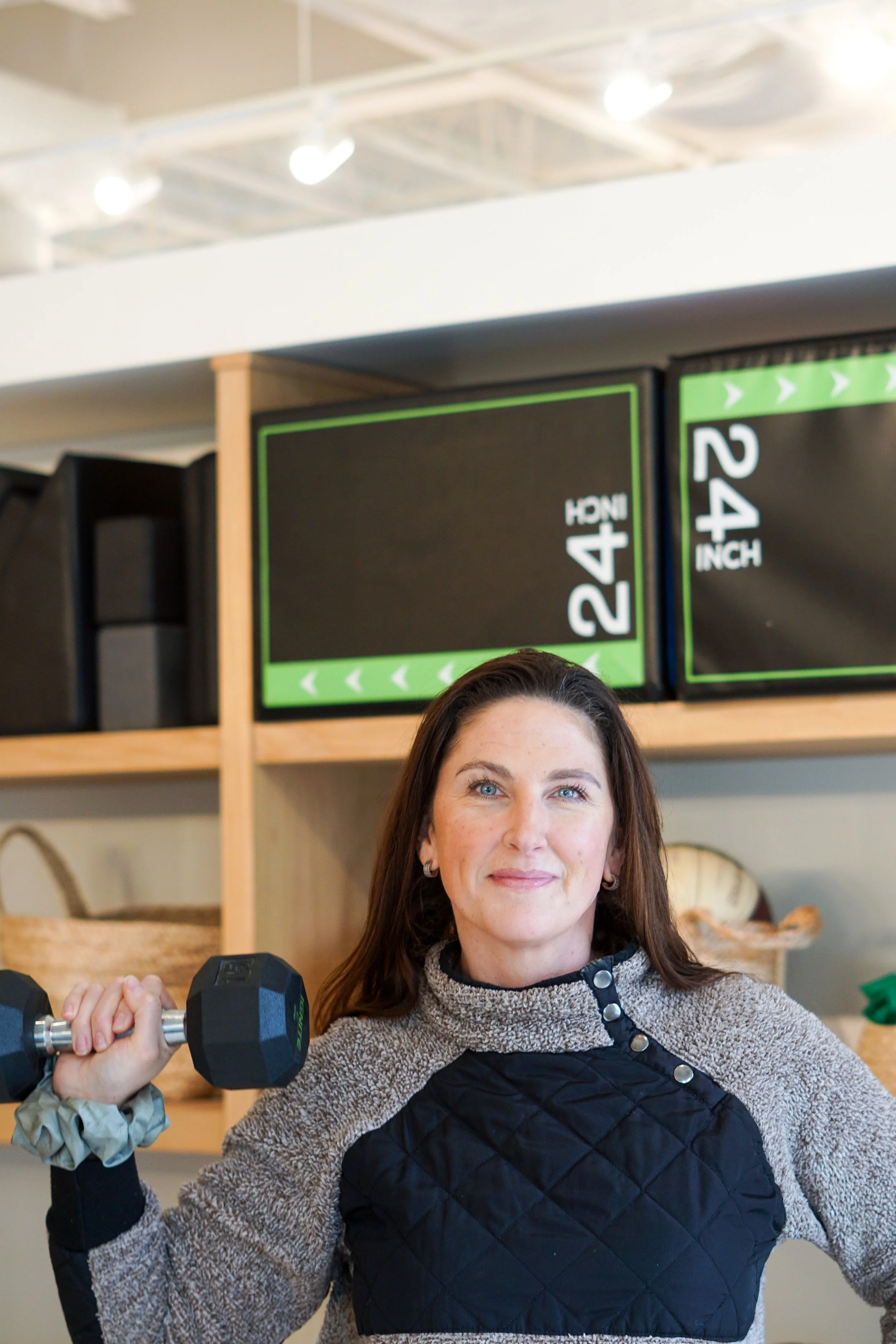 Woman holding a dumbbell in a gym or fitness studio, with height measurement signs on the shelf behind her.