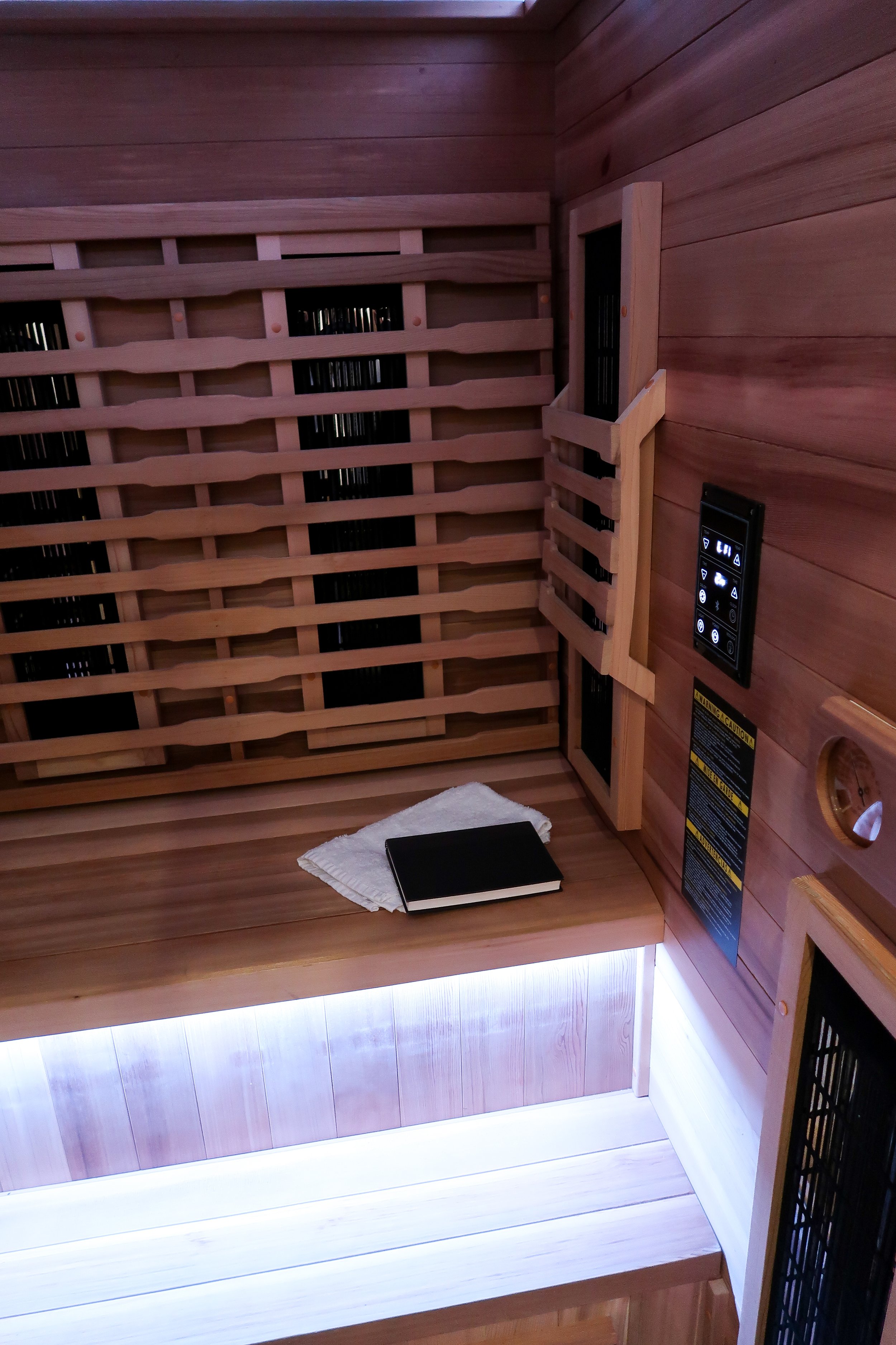 Inside a wooden infrared sauna with a heated bench, a white towel, and a closed black book. Wooden walls with an adjustable vent and a control panel.