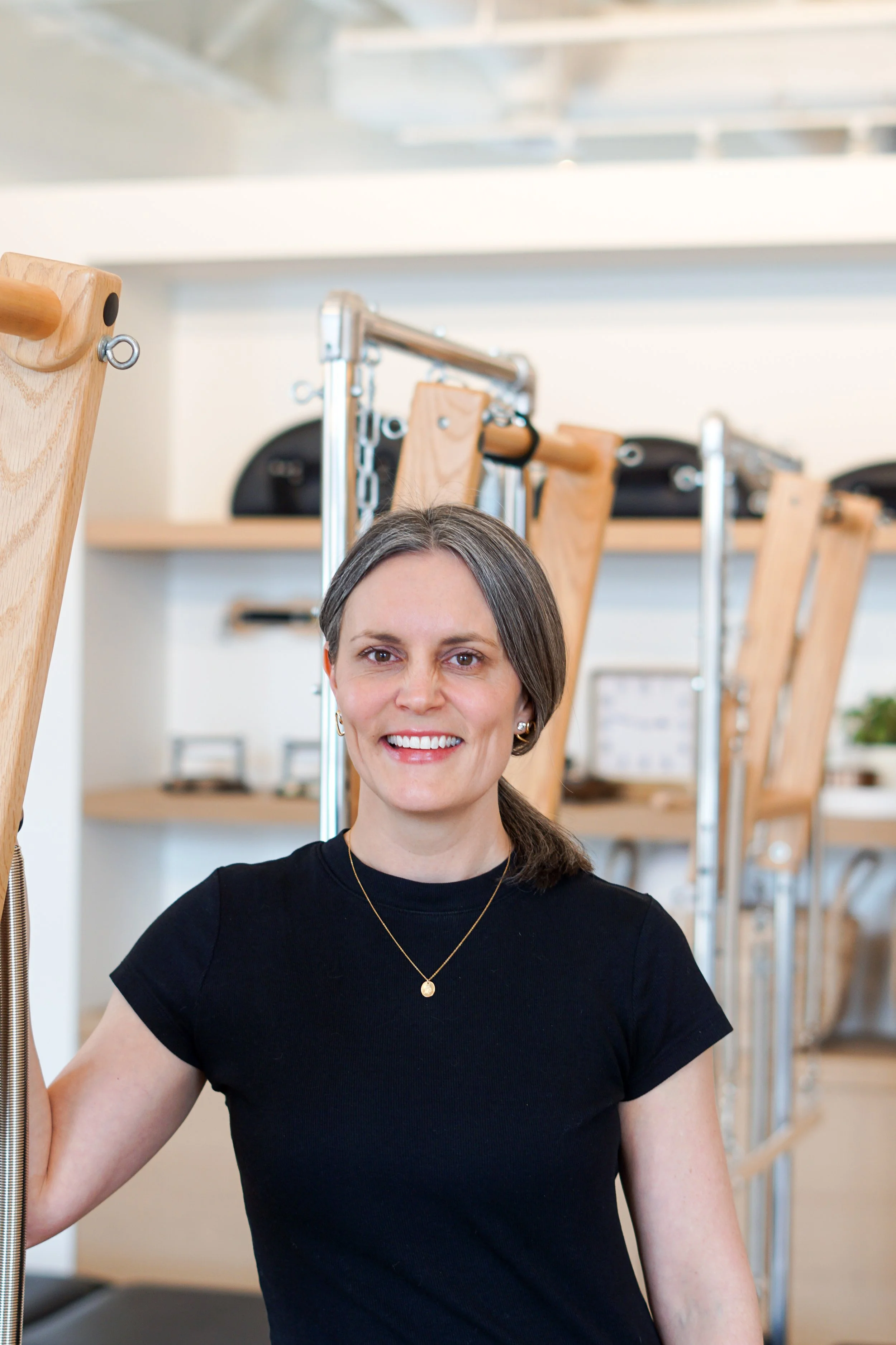 A smiling woman with gray hair tied back, wearing a black t-shirt and a gold necklace, standing in a fitness or pilates studio with wooden and metal exercise equipment in the background.