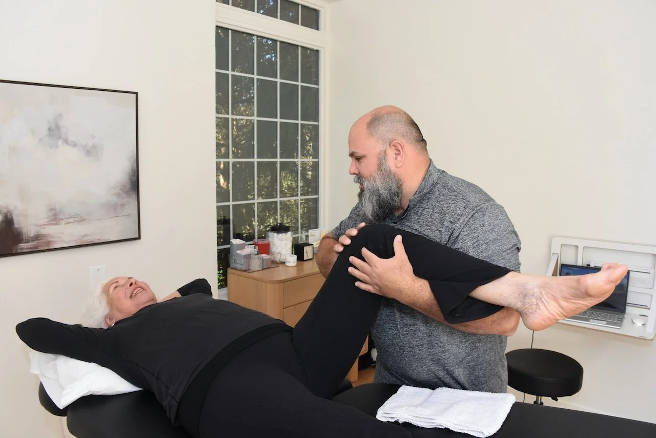 A healthcare professional aids an elderly woman in a physical therapy exercise, lifting her leg while she lies on a treatment table, smiling.
