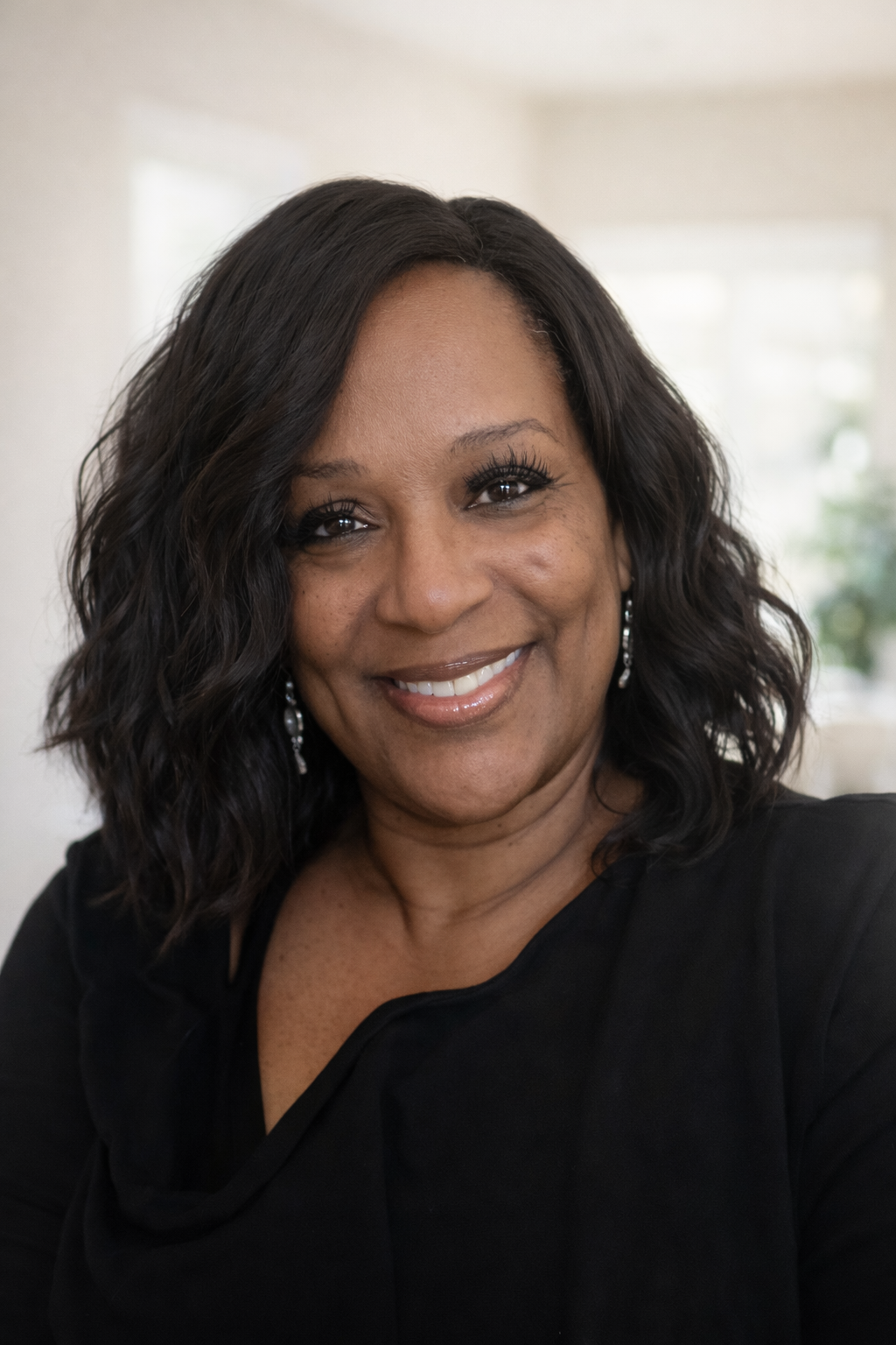 A smiling woman with black wavy hair is wearing a black top and silver earrings; she is indoors with a softly lit background.