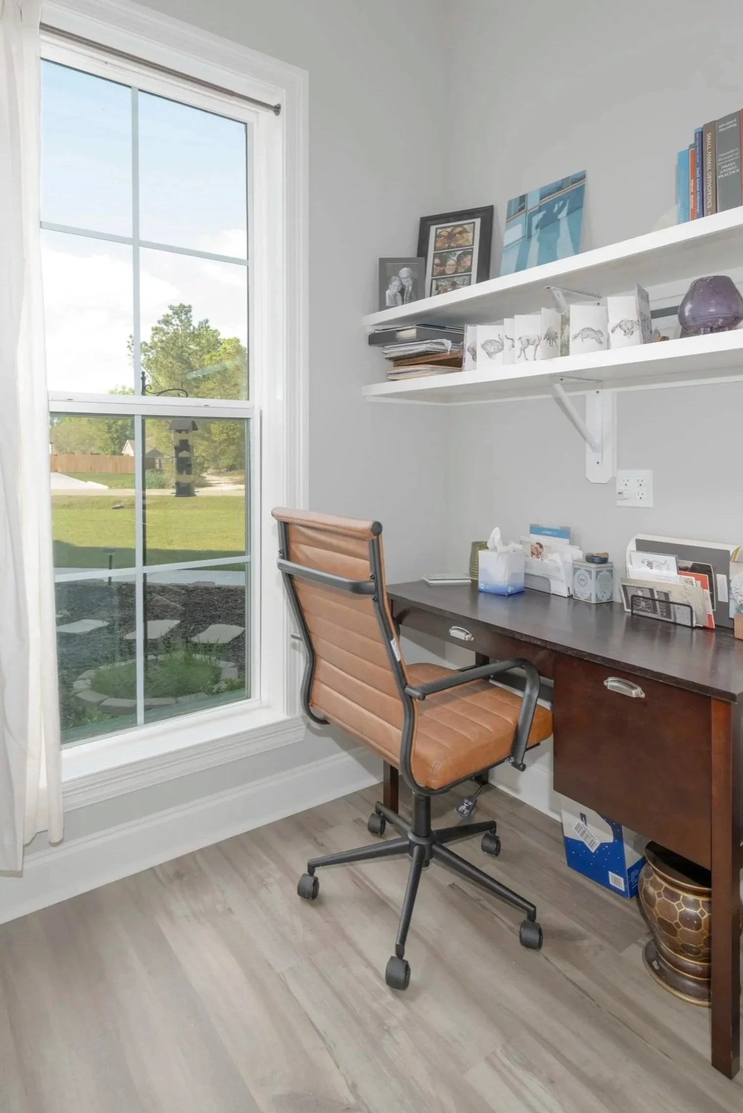 A home office with a large window showing trees and a birdhouse outside. Inside, there is a brown swivel office chair in front of a dark wood desk with various items, including a tissue box, a stack of papers, and framed photos. White shelves above the desk hold books, framed pictures, and decorative objects, including a purple vase. The floor is light-colored wood.