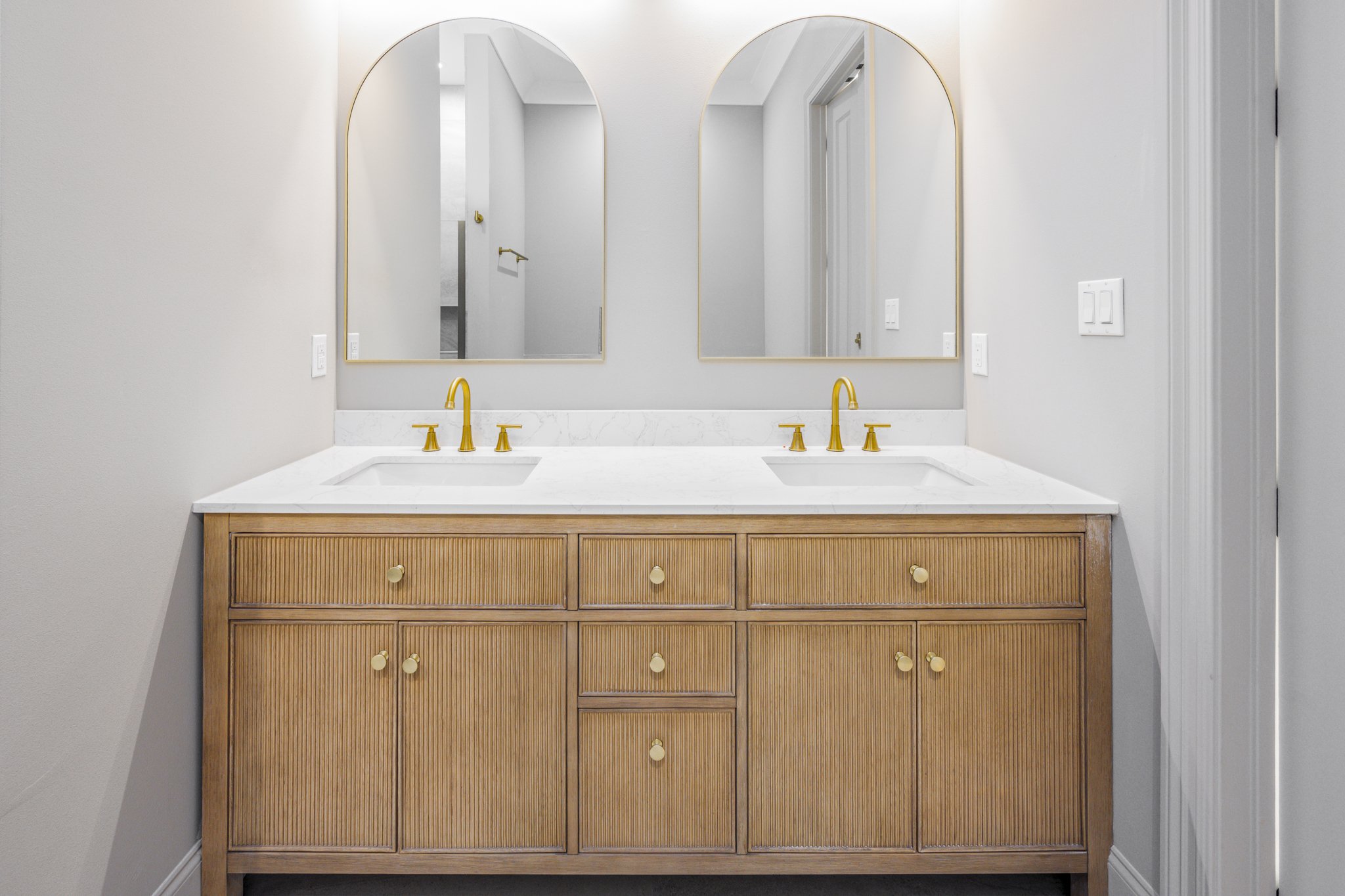 A bathroom vanity with a white marble countertop, two sinks, and gold faucets, with a large mirror above it and a wooden cabinet with multiple drawers.