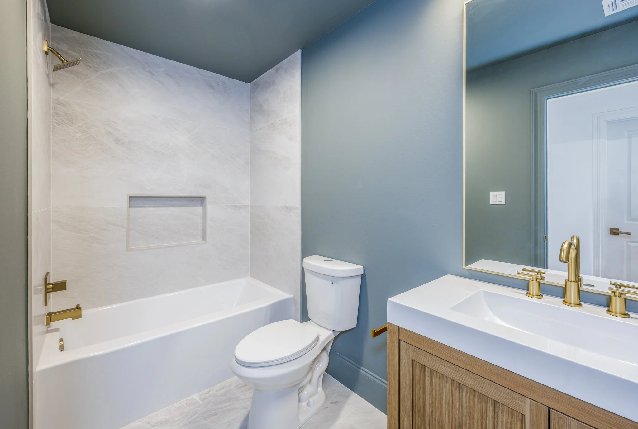 A modern bathroom with a bathtub, toilet, and sink with gold fixtures, light blue walls, a large mirror, and a window with a reflection of a door.