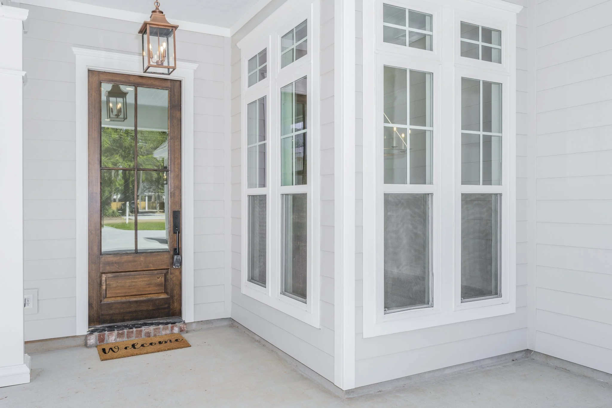 Front porch of a house with a wooden door, a doormat that says 'welcome,' and a white screen door with a lantern-style light fixture hanging above.