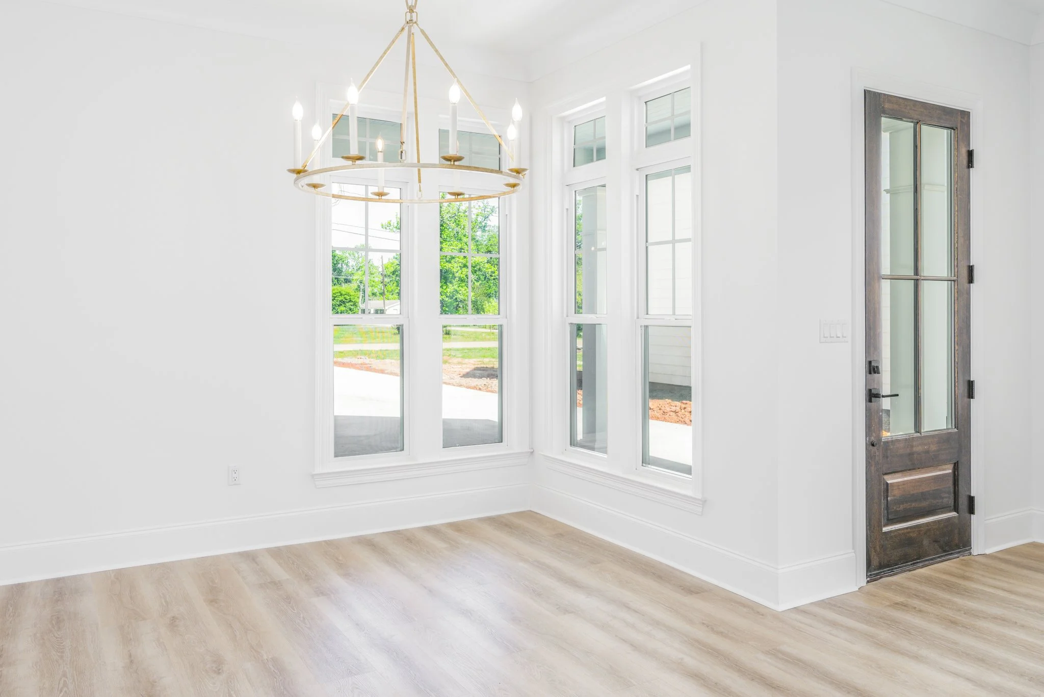 Empty living room with large windows, light wood flooring, a modern circular chandelier, and a wooden front door.