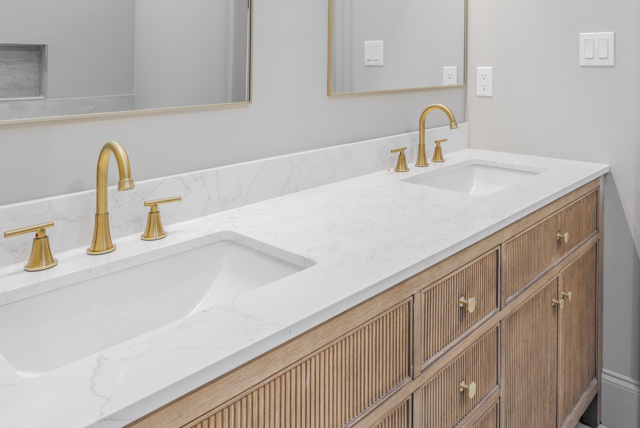 Bathroom vanity with a white marble countertop, two sinks, and six gold faucets, with a wooden cabinet below, two mirrors above, and electrical outlets and switches on the wall.