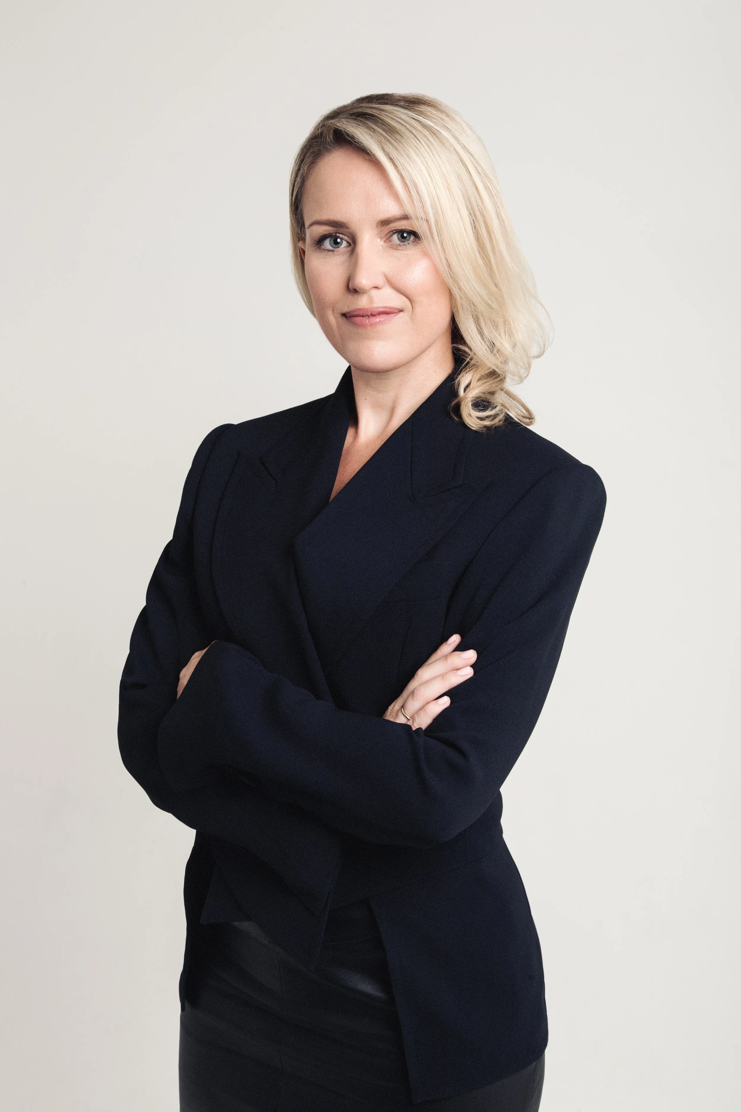 Portrait of a professional woman with blonde hair, wearing a black blazer, with arms crossed, standing against a plain light background.