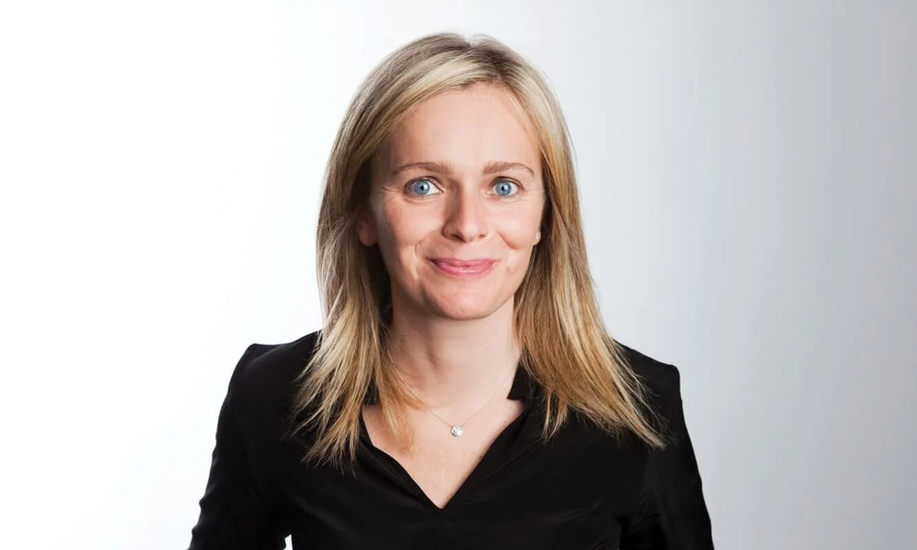 A woman with blonde hair, blue eyes, wearing a black blazer and a necklace, smiling at the camera against a plain background.