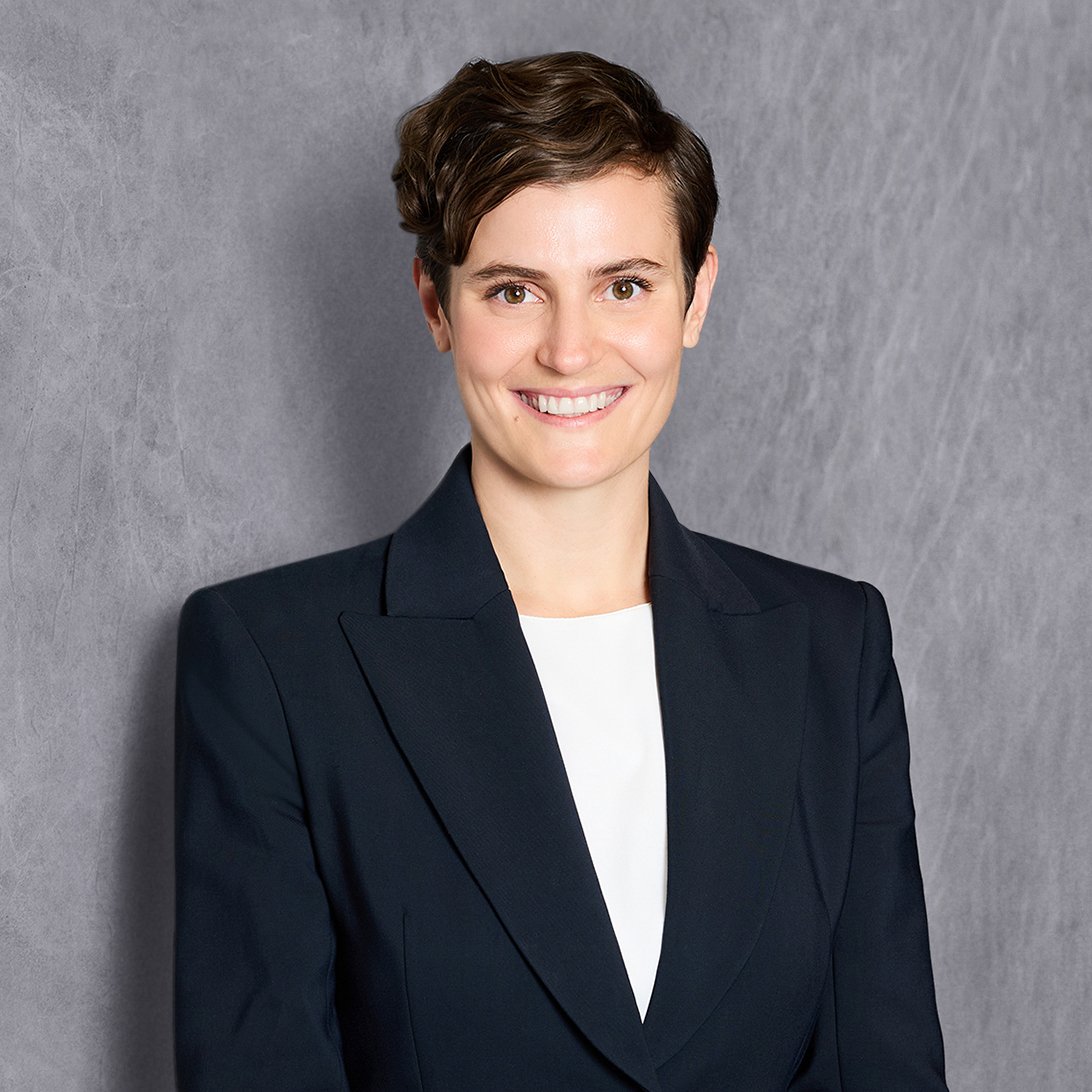 A woman with short brown hair wearing a black blazer is smiling against a textured gray wall.