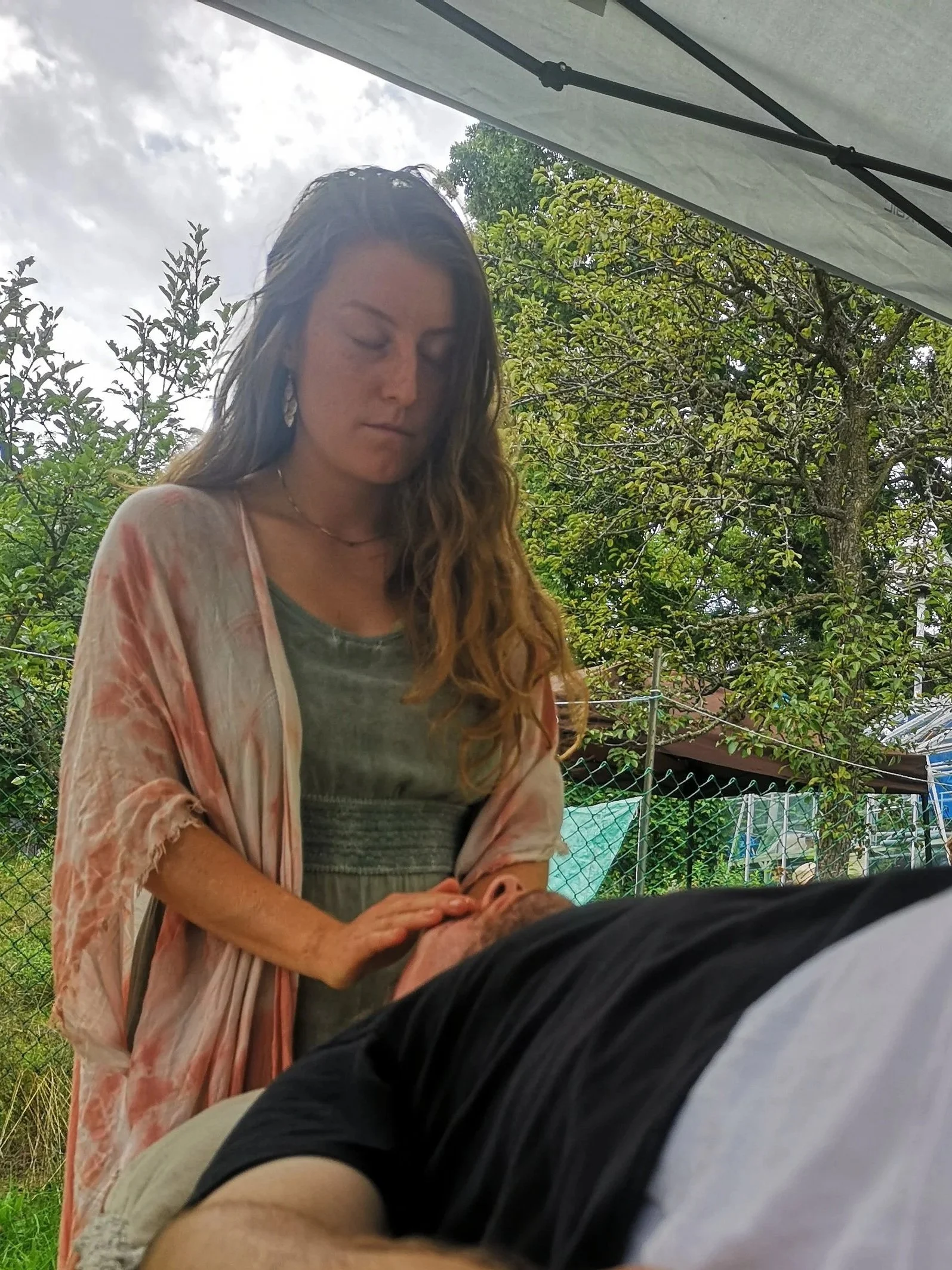 A woman with long wavy hair is giving a massage outdoors under a tent, with trees and a cloudy sky in the background.