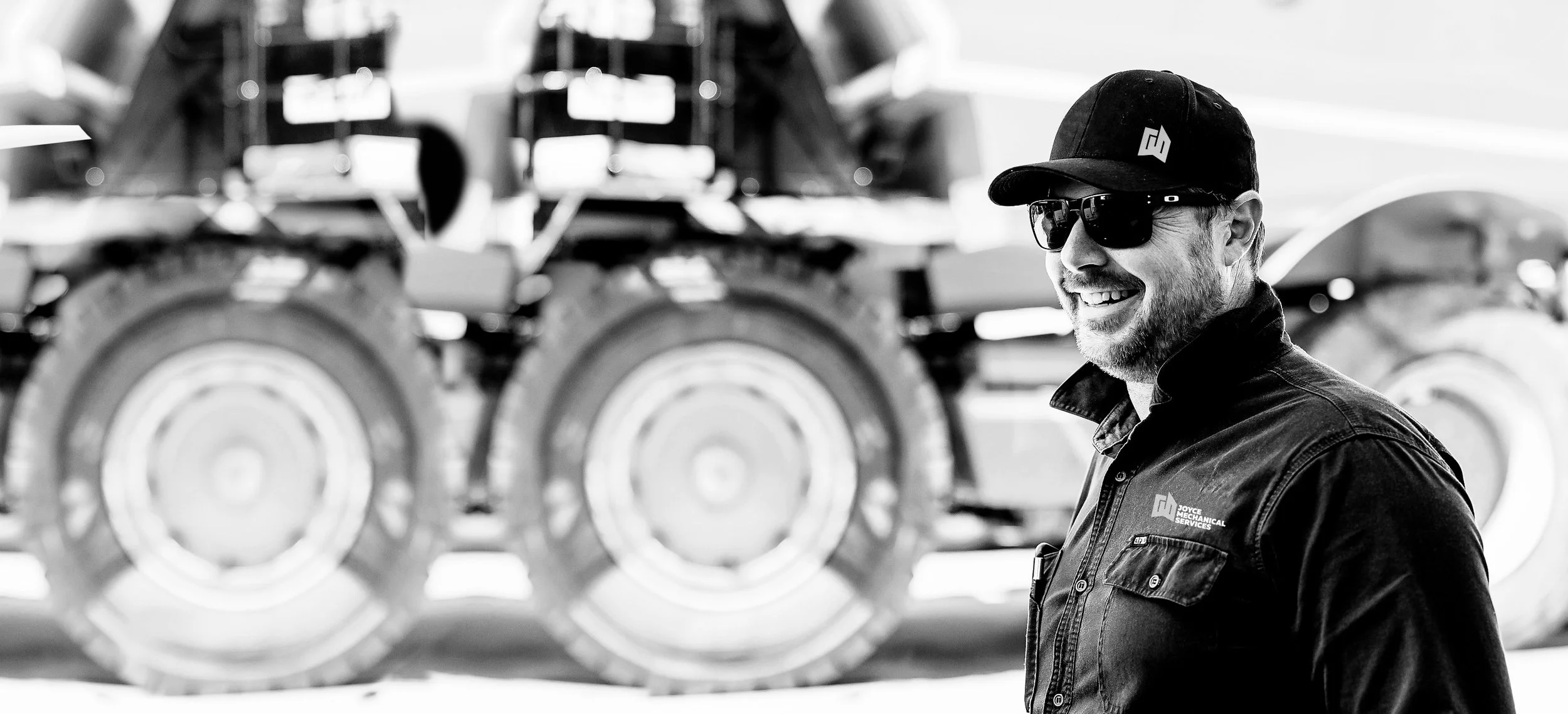 A man wearing sunglasses, a cap, and a jacket smiling in front of a large piece of machinery with multiple wheels.