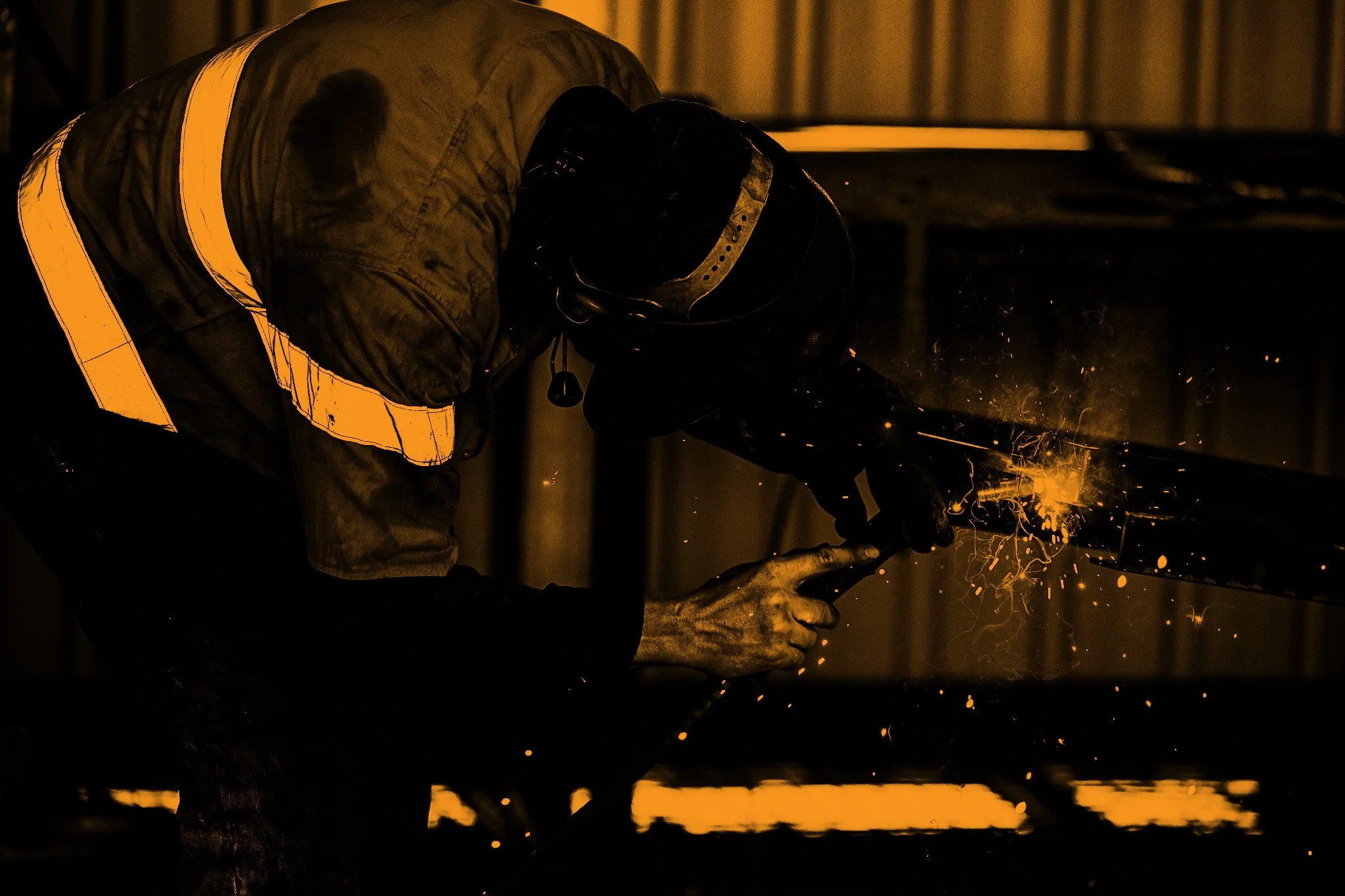 A worker welding in a dark industrial setting, with sparks flying from the weld.