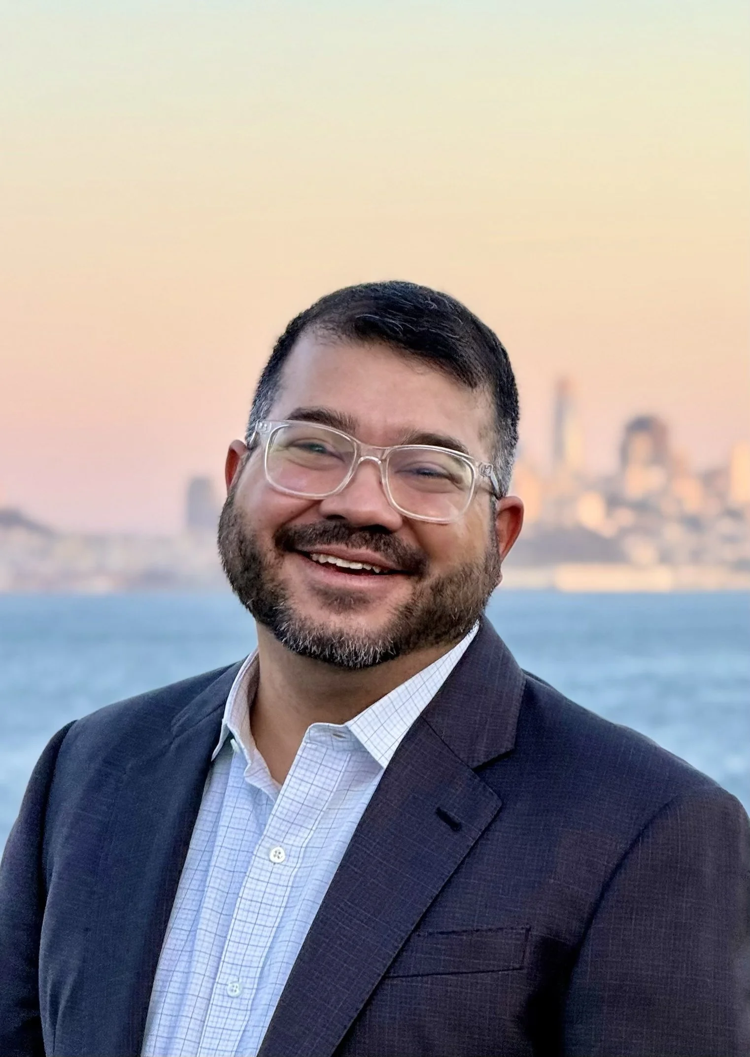 A smiling man in a suit and glasses standing outdoors by the water with a city skyline in the background at sunset.
