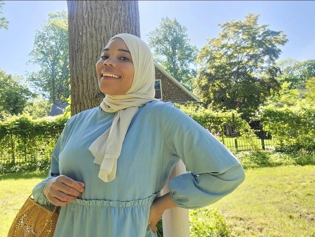 A woman smiling outdoors in a sunny backyard with green trees, a large tree trunk, a house with a stone facade, and a black fence in the background. She is wearing a light-colored hijab and a light blue dress.
