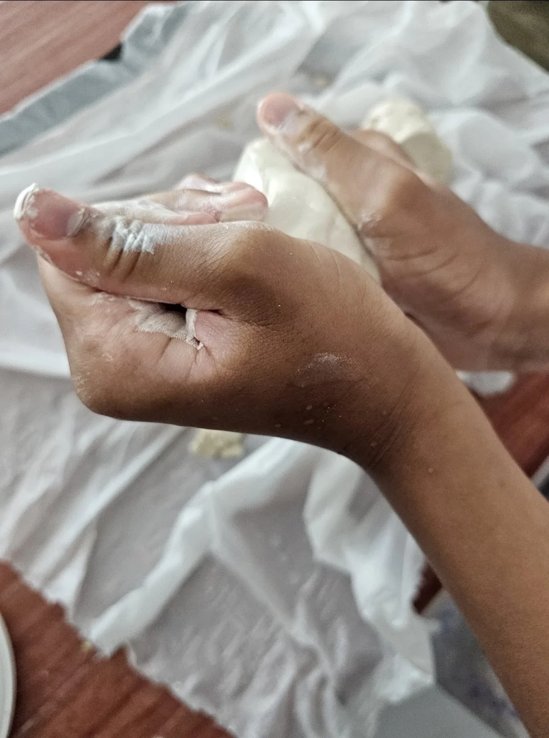 Person hands covered with white cream or lotion, resting on a white cloth with a wooden surface in the background.