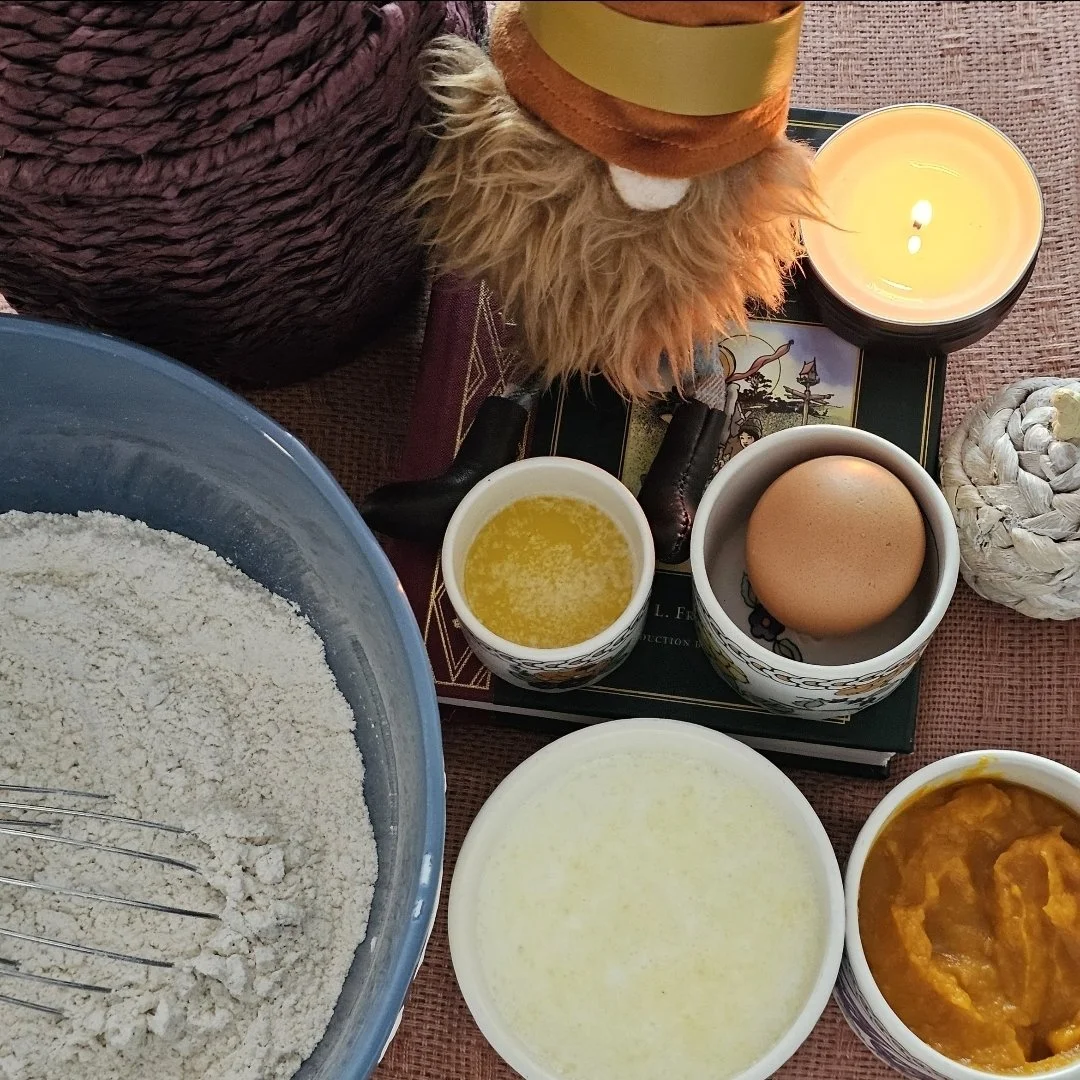 A baking setup with ingredients and decorations including flour, egg, butter, pumpkin puree, a lit candle, a small plush lion wearing a hat, and baking utensils on a textured table surface.