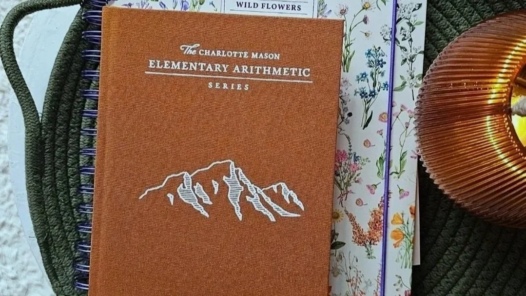 An orange textbook titled "The Charlotte Mason Elementary Arithmetic Series" on a woven dark green circular surface, with a floral-patterned book or paper underneath and part of a copper-colored decorative object visible.