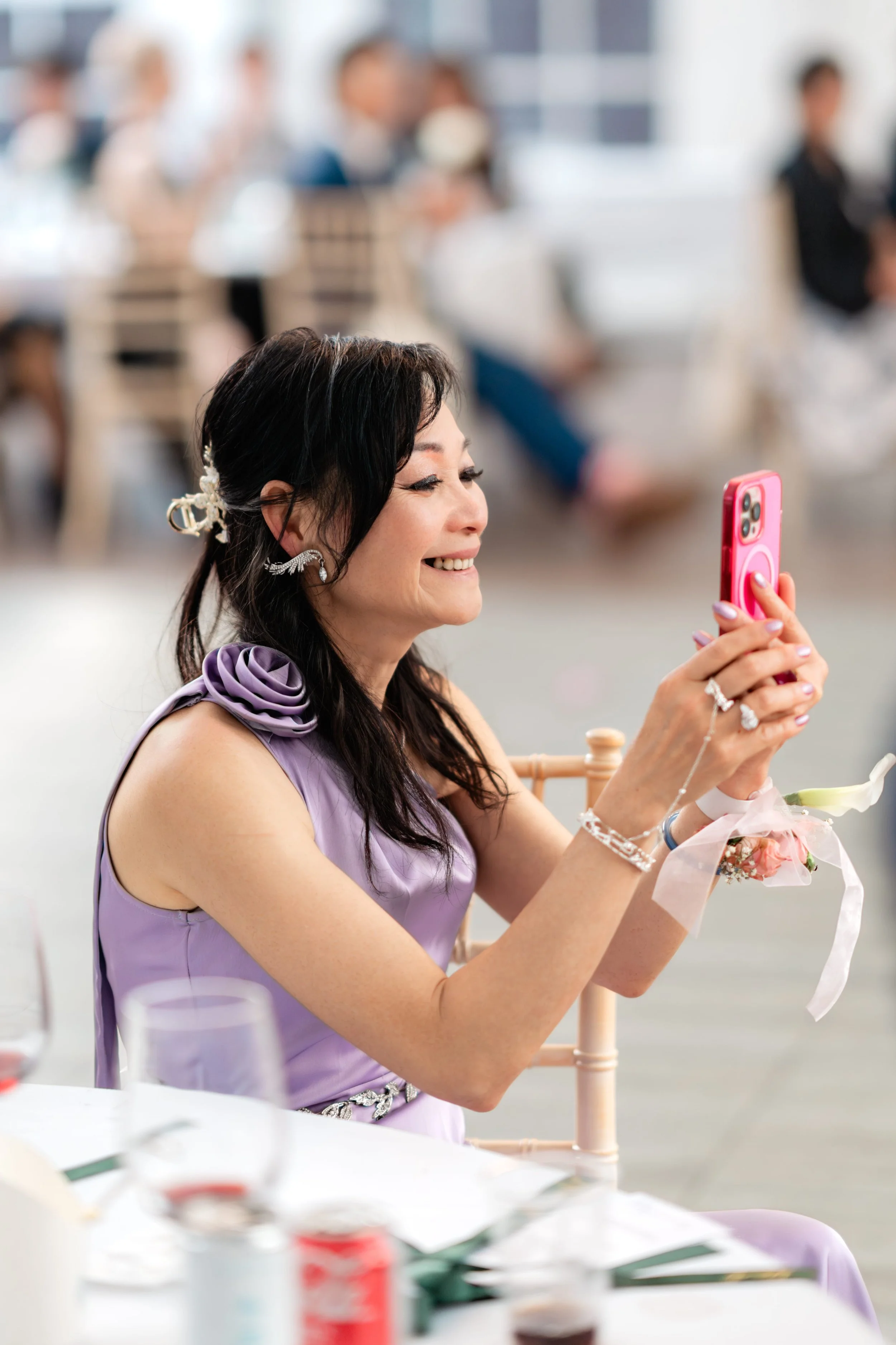 A woman in a purple dress sitting at a table, taking a selfie with her phone at a social event or celebration.