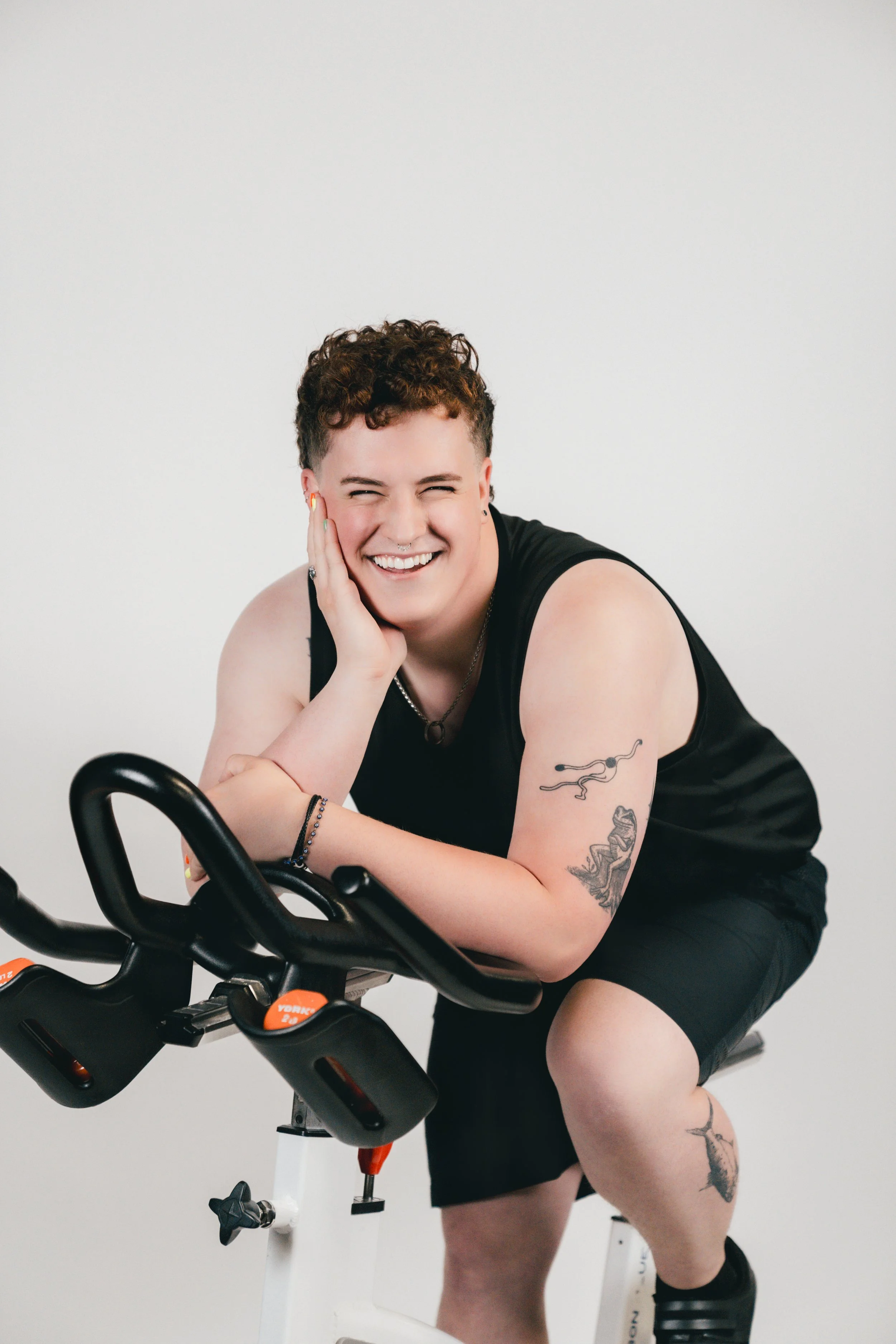 A person with short curly hair and tattoos, wearing a black sleeveless shirt, sitting on a stationary exercise bike, smiling and resting their face on their hand against a plain white background.