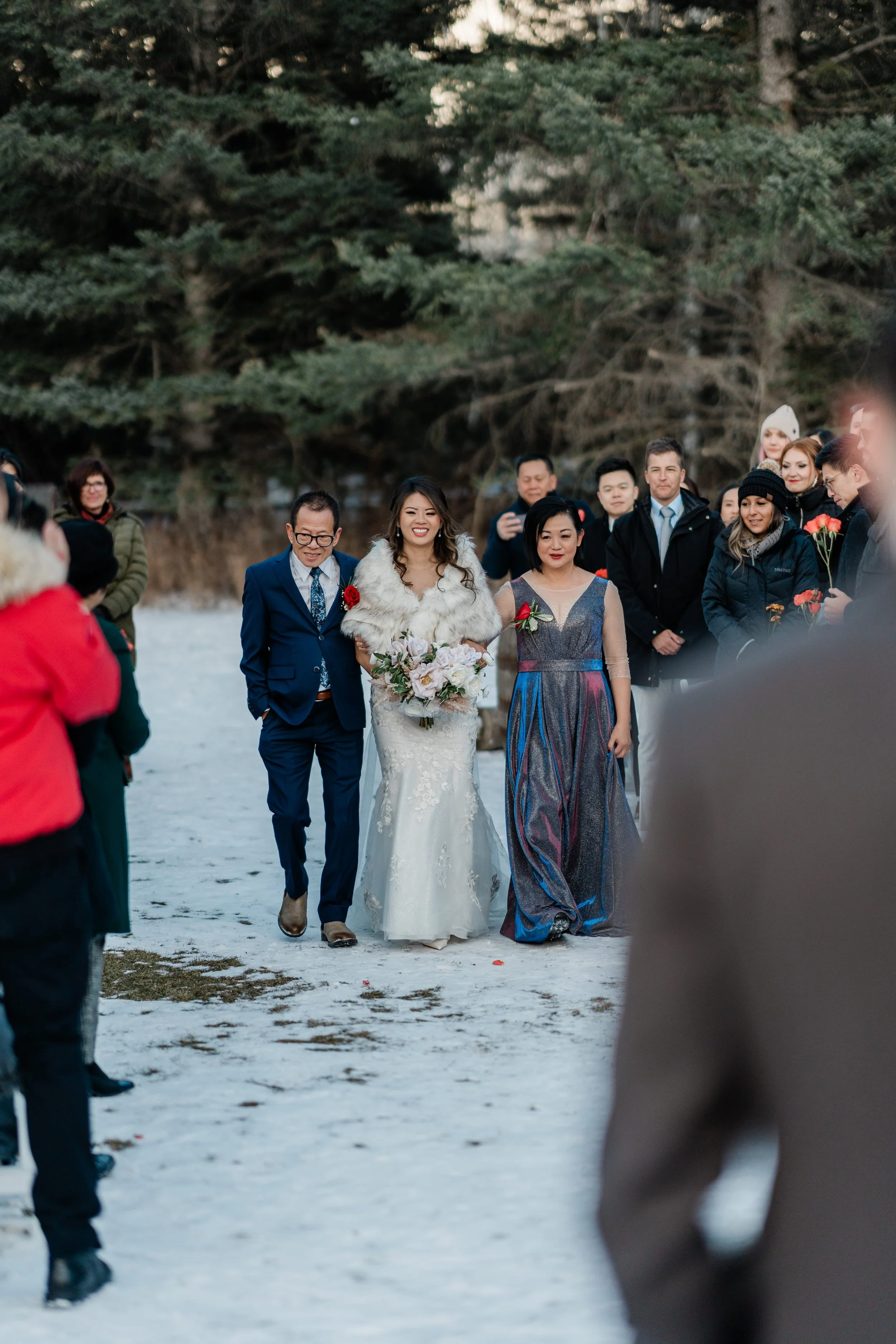 A bride in a white dress and fur stole walking outdoors on snow-covered ground, flanked by people dressed in formal and winter clothing, with trees in the background.