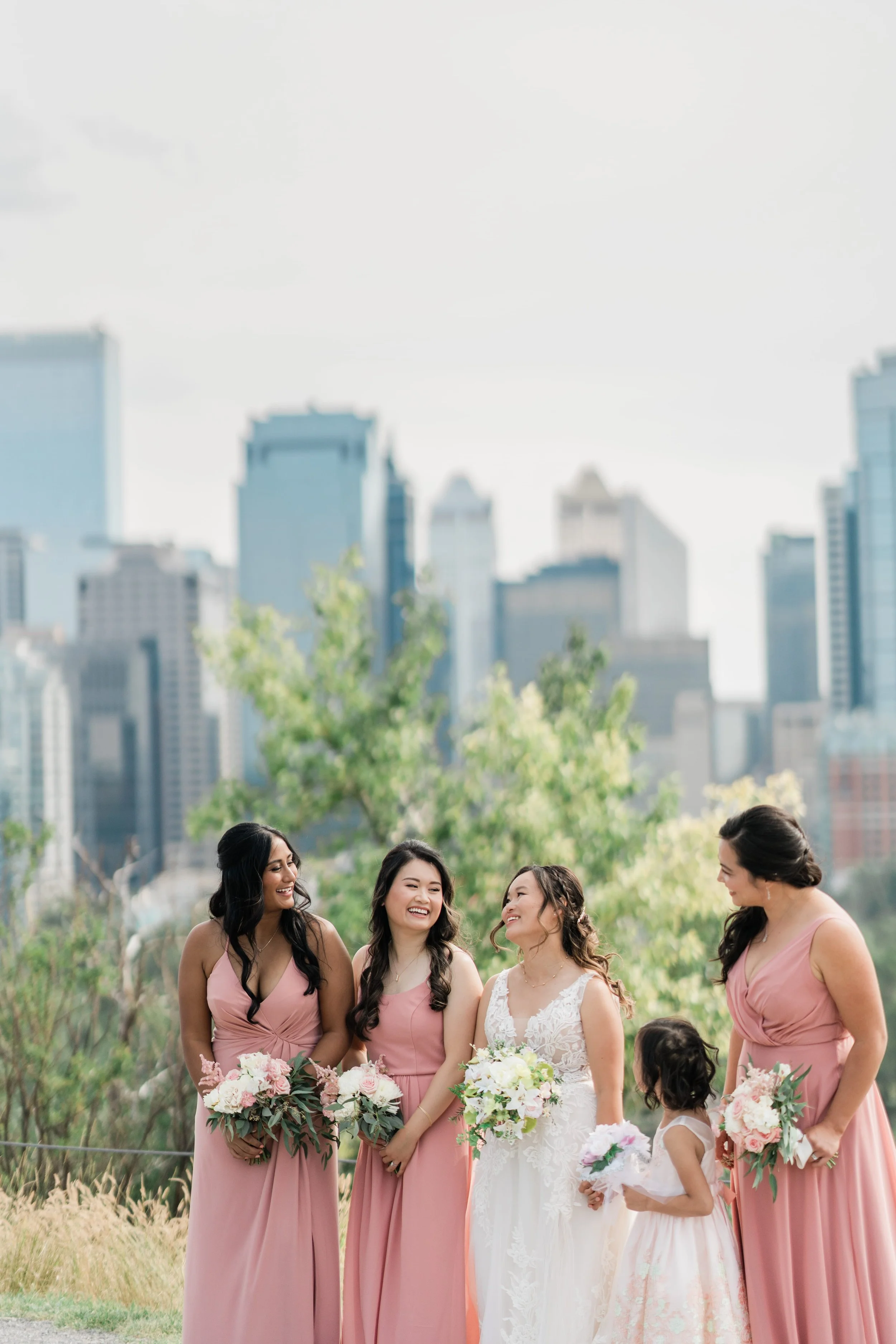 A bride and four bridesmaids standing outdoors, dressed in pastel pink and white gowns, holding bouquets, and smiling at each other with a city skyline and trees in the background.
