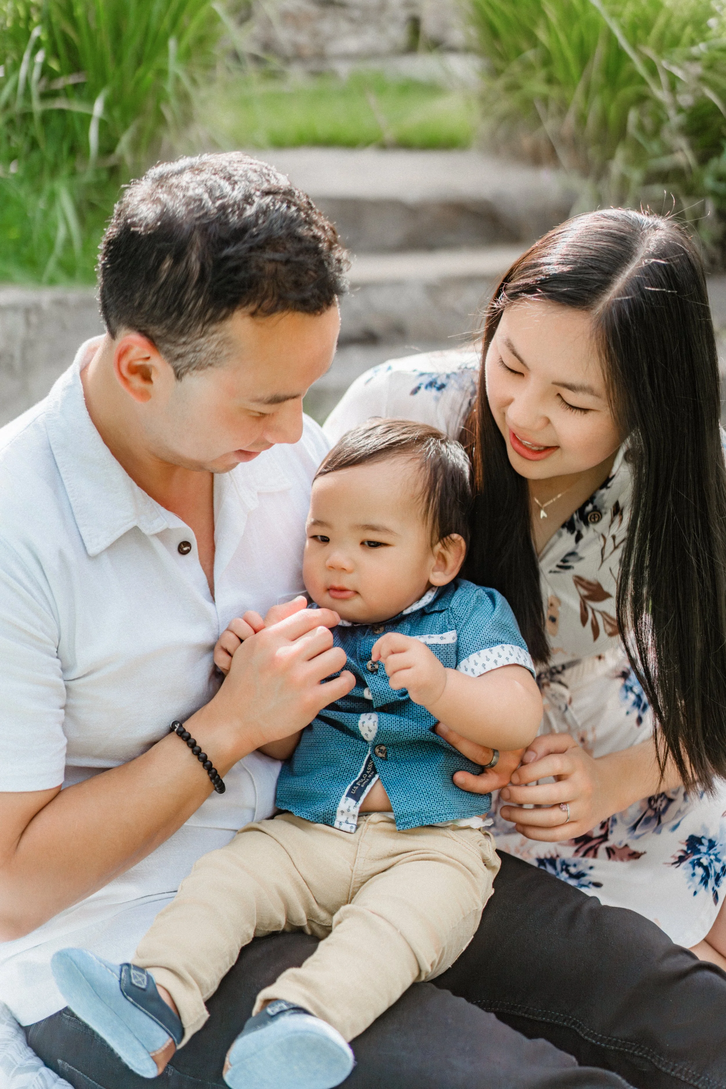 A family of three, including a man, woman, and young child, sitting outdoors amidst greenery, enjoying a moment together.