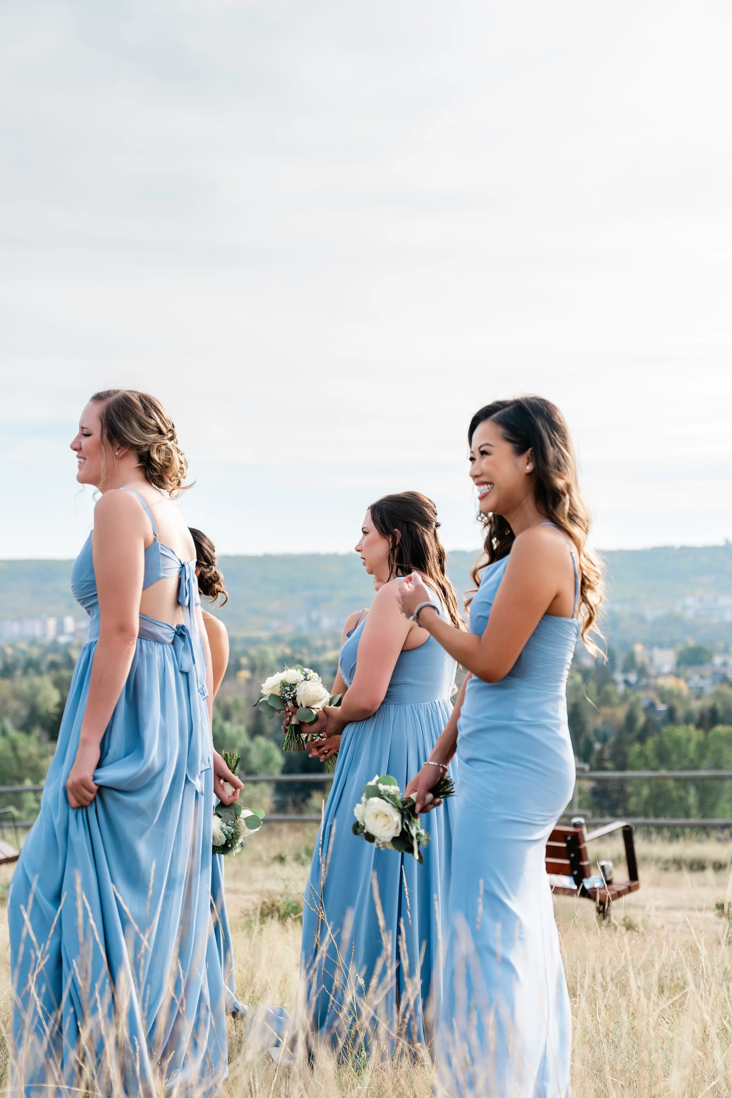 Group of women in blue dresses holding bouquets, standing outdoors in a grassy field, smiling and chatting.