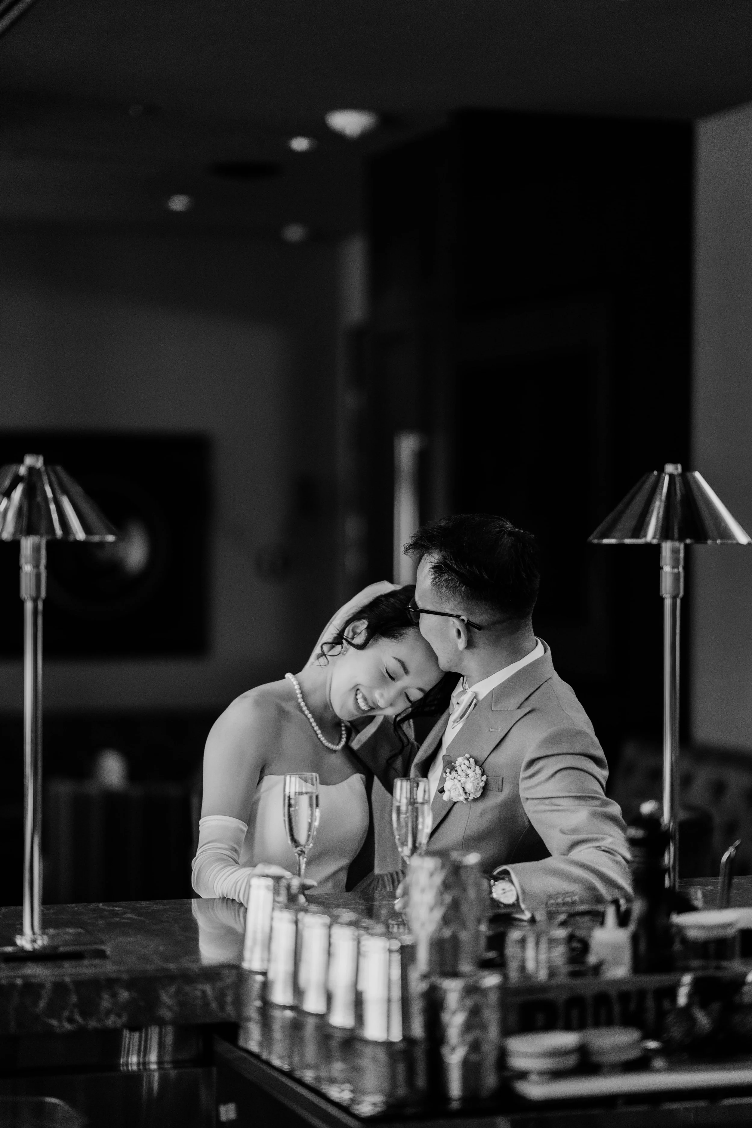 A black and white photograph of a newlywed couple sharing an intimate moment at a celebration, with champagne glasses on a table in front of them.