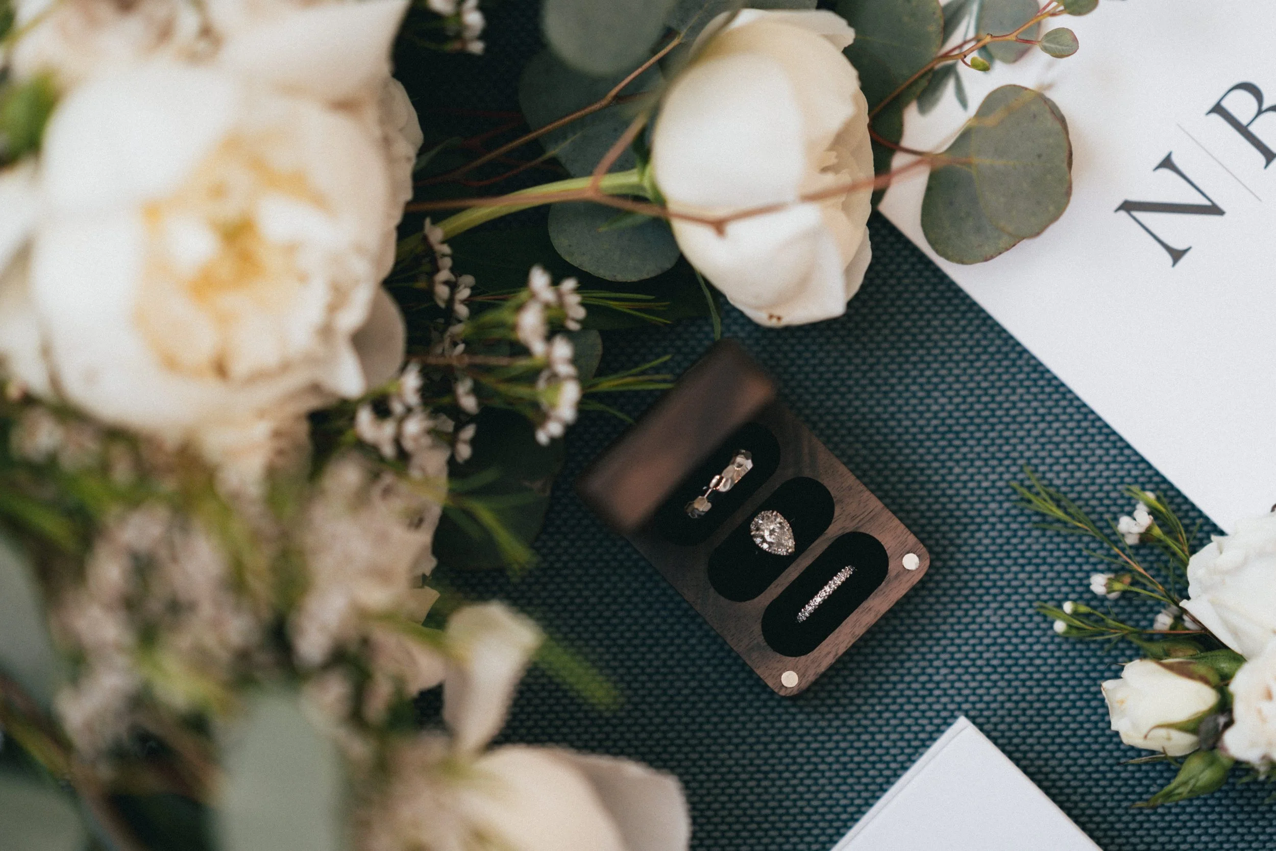 Close-up of a jewelry display with rings and stud earrings, surrounded by white flowers and greenery on a dark textured surface.