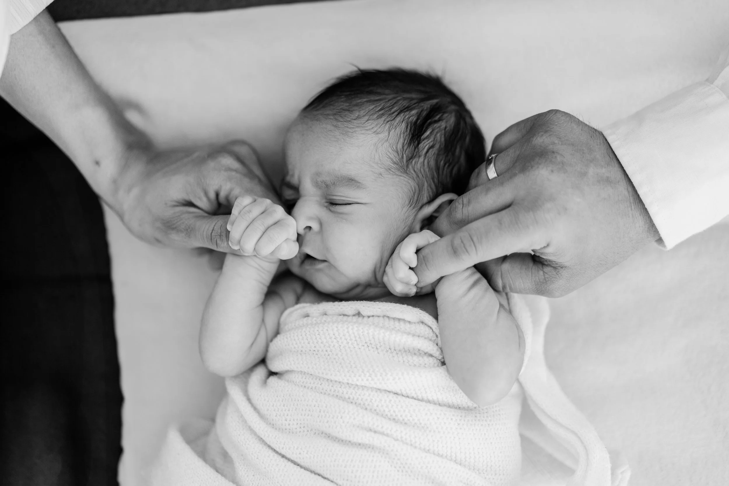 A newborn baby wrapped in a blanket, sleeping peacefully while being held and gently touched by two adults' hands.