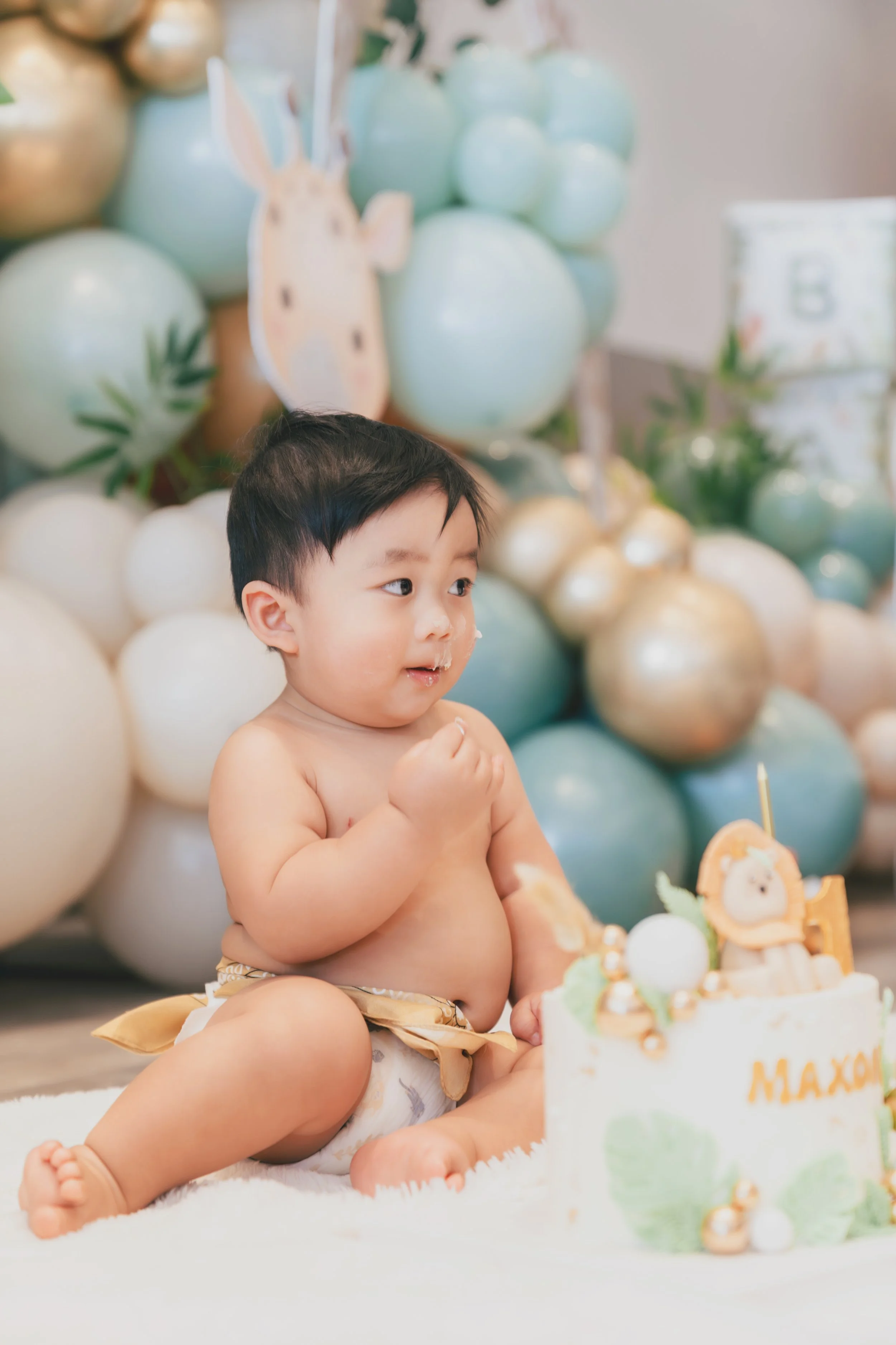 A young child sitting on a white rug during a birthday celebration, with a decorated birthday cake in front and a backdrop of pastel-colored balloons and a bunny-themed decoration.