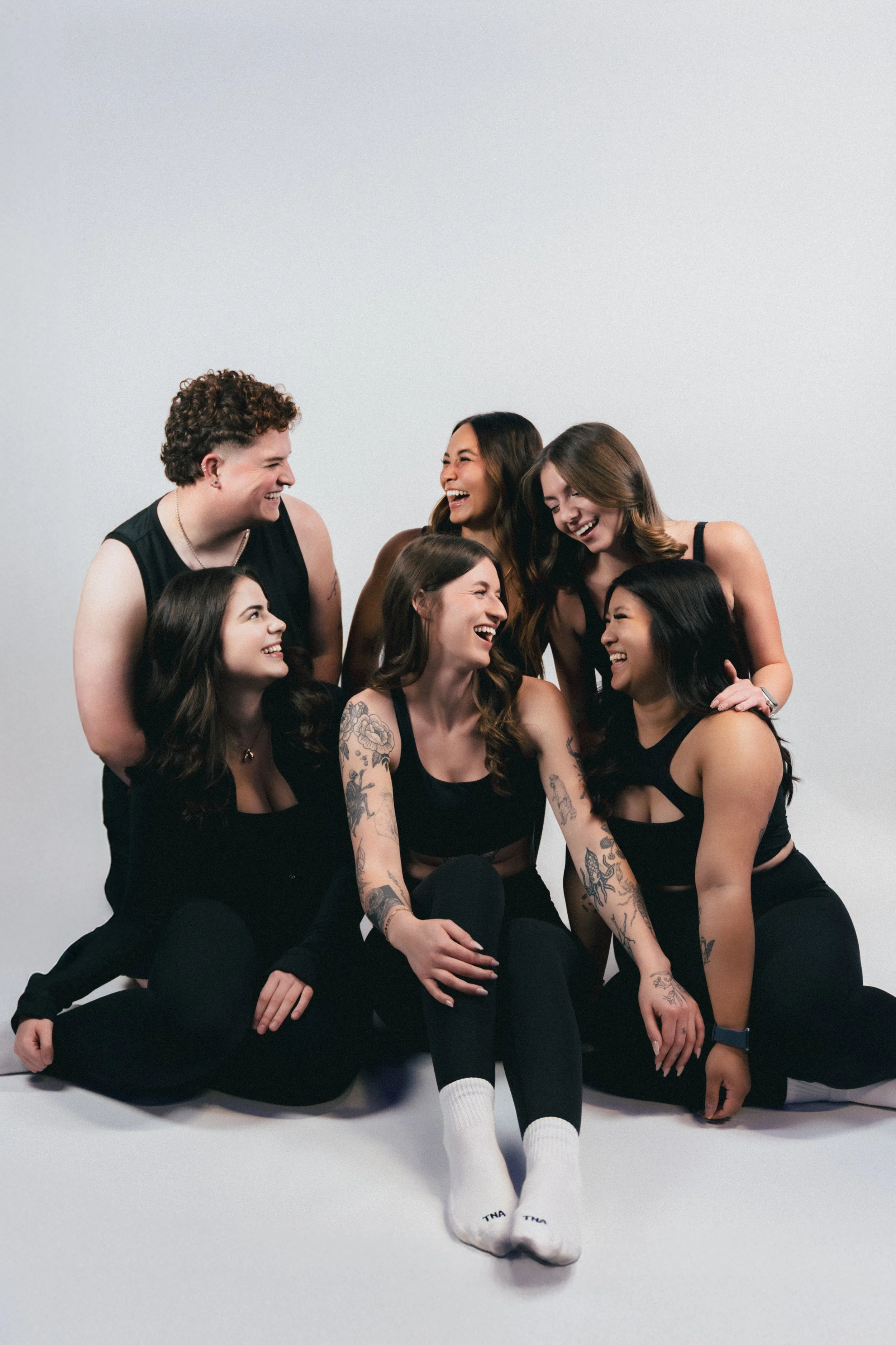 Group of six women laughing and smiling together in a studio against a plain gray background.