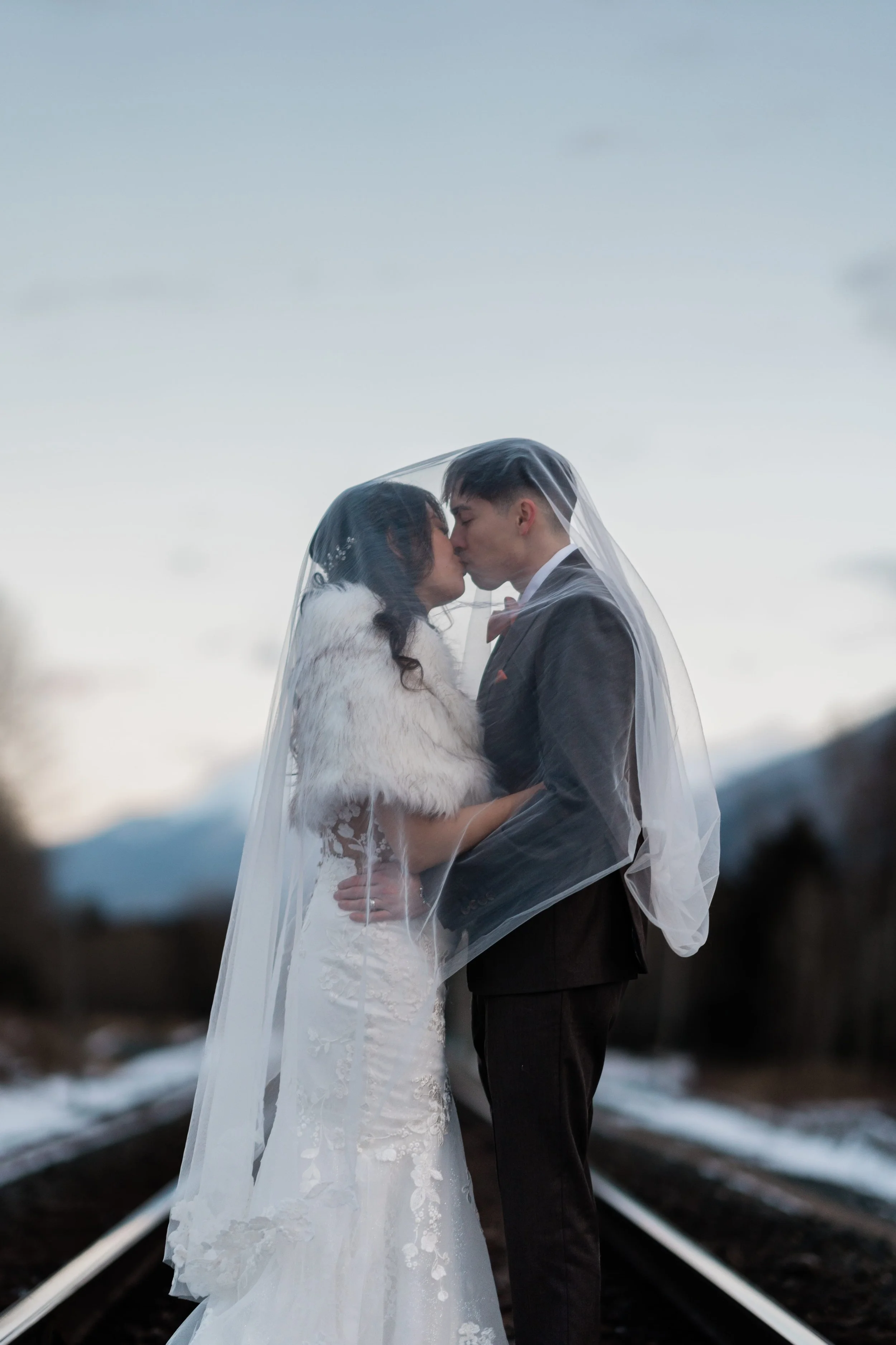 A bride and groom sharing a kiss on train tracks with mountains in the background, under a partly cloudy sky
