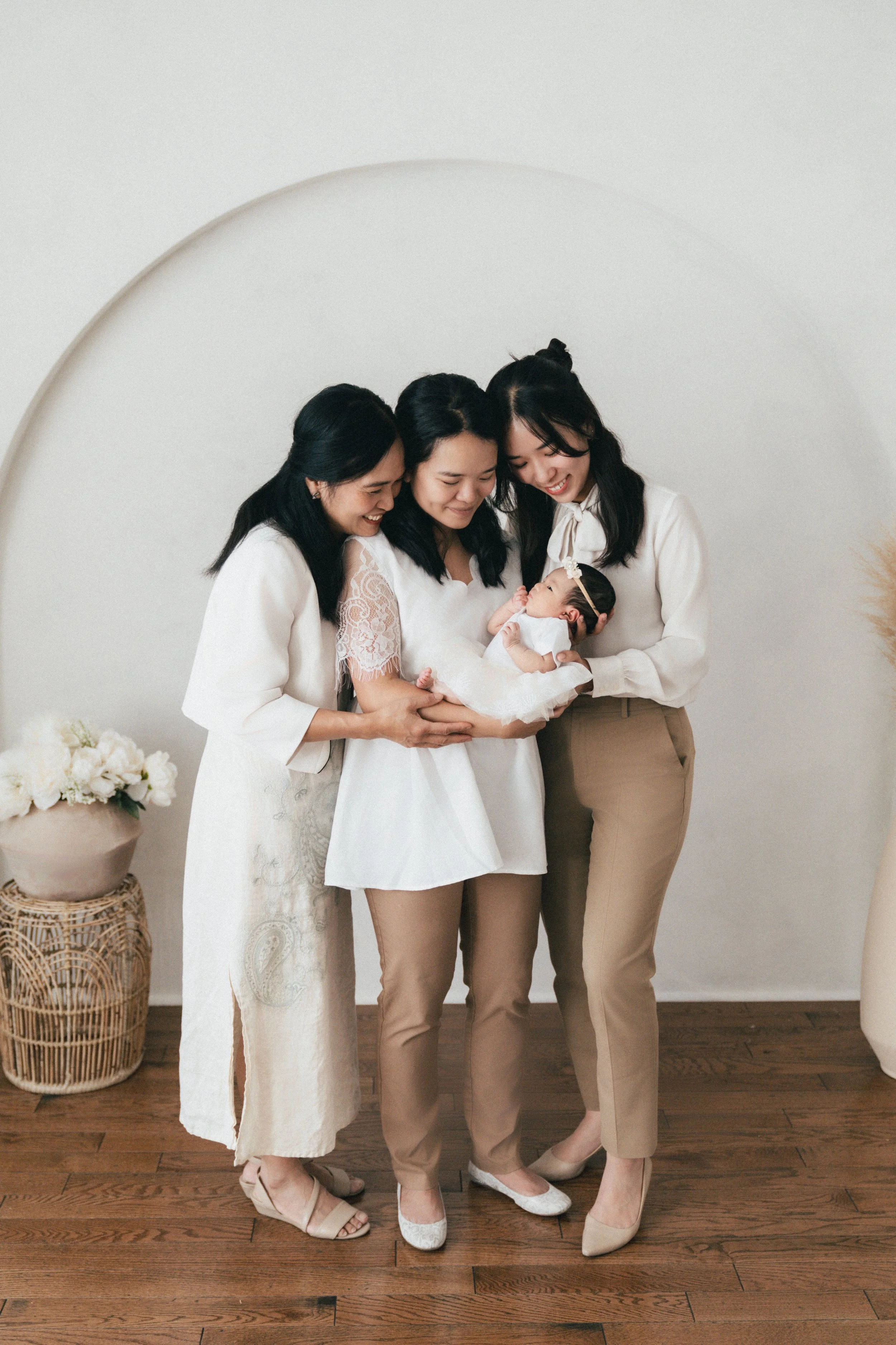 Four women, dressed in neutral tones, gather closely and smile while holding a baby girl dressed in white with a headband, against a simple white wall and wooden floor.