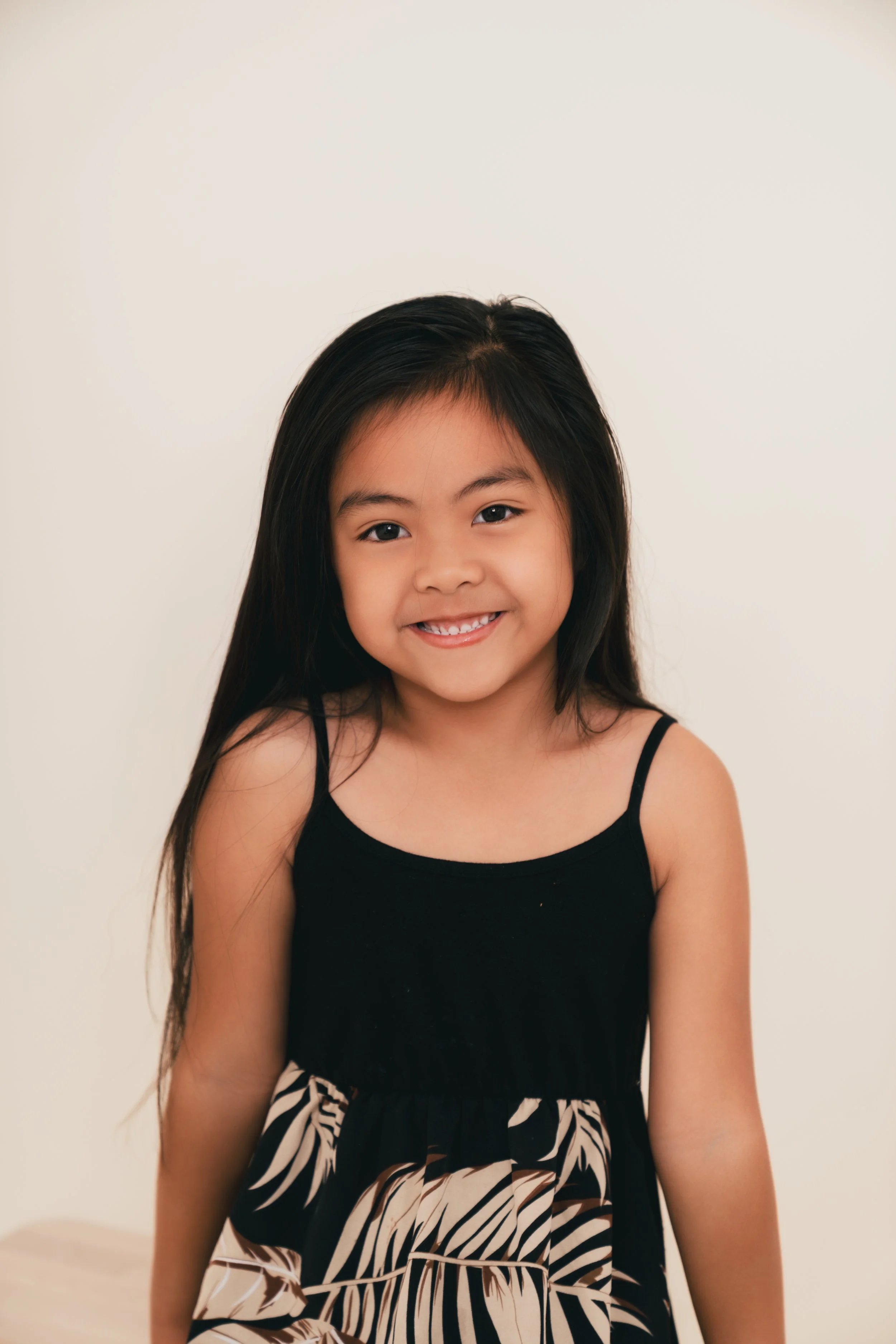 A young girl with long black hair, smiling, wearing a black dress with a beige and black leaf pattern.