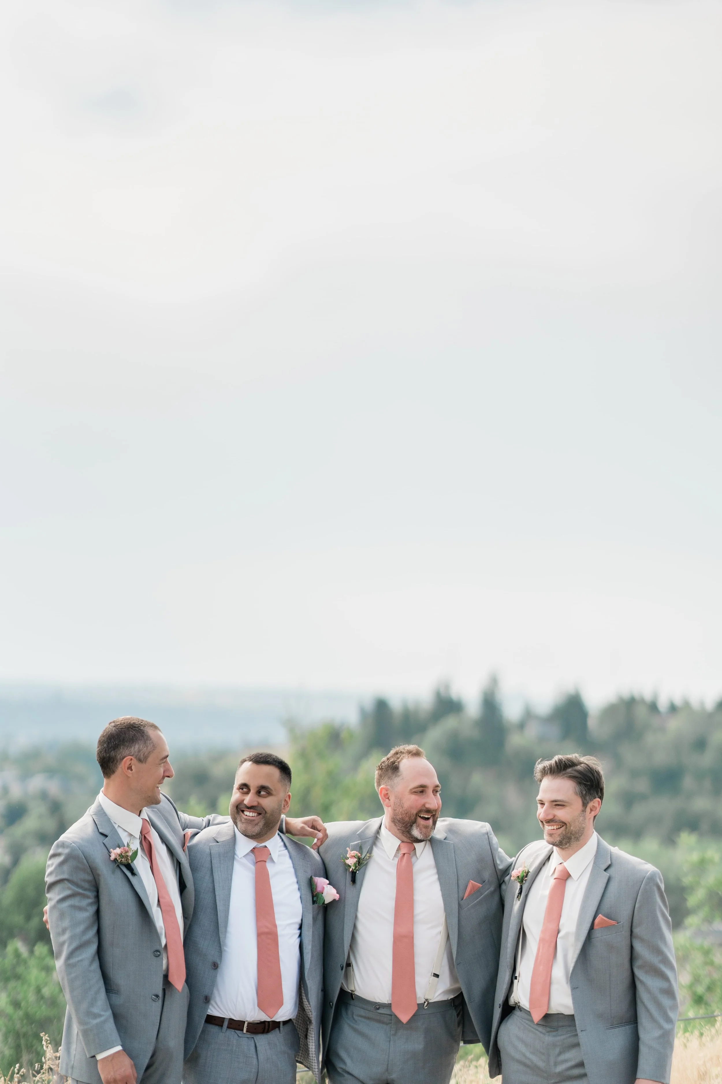 Four men in gray suits with pink ties and boutonnieres, standing outdoors and smiling with their arms around each other, on a scenic hilltop with trees and hills in the background.