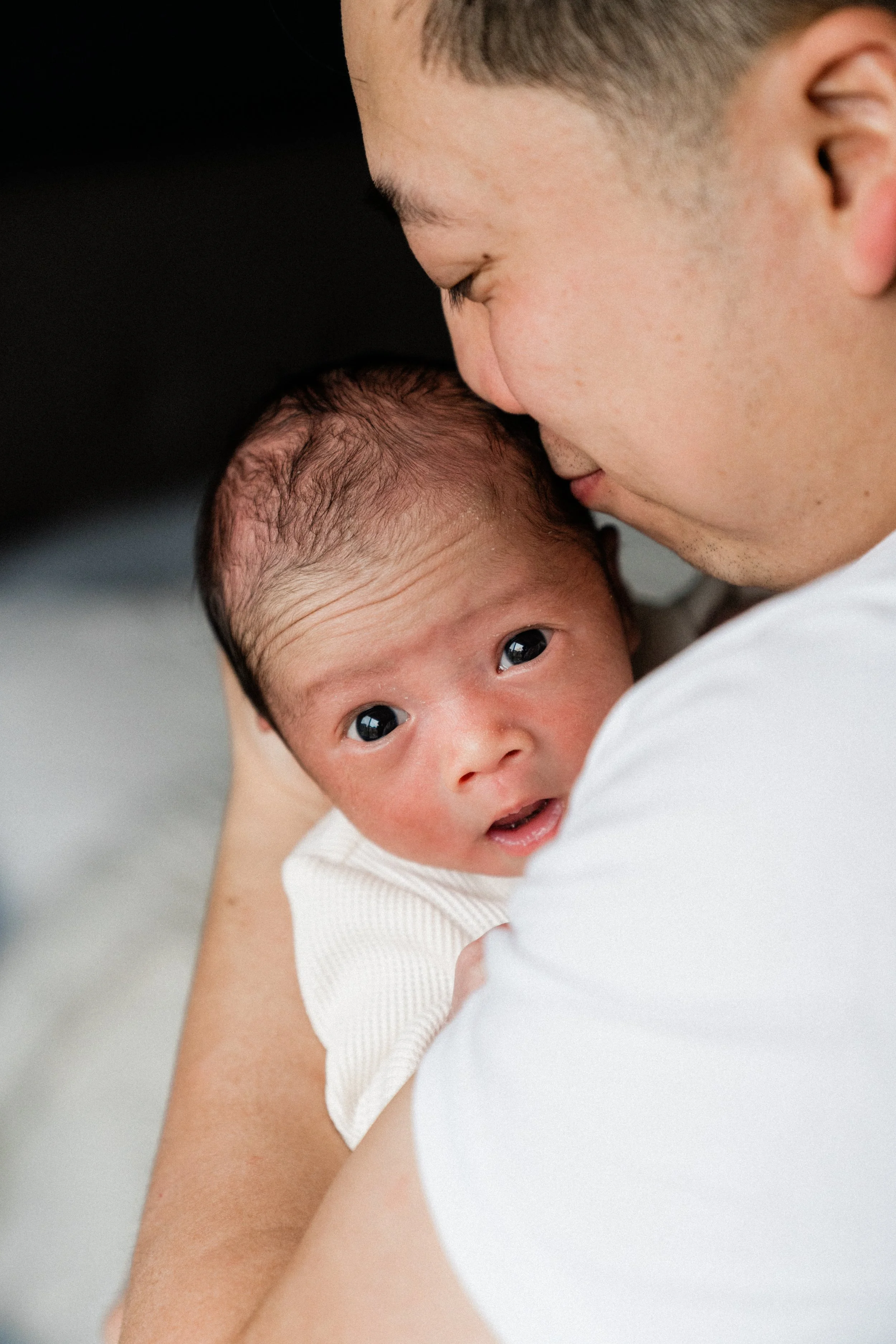 A man holding a newborn baby close, with the man's face gently touching the baby's head. The baby is looking directly at the camera with wide, curious eyes.