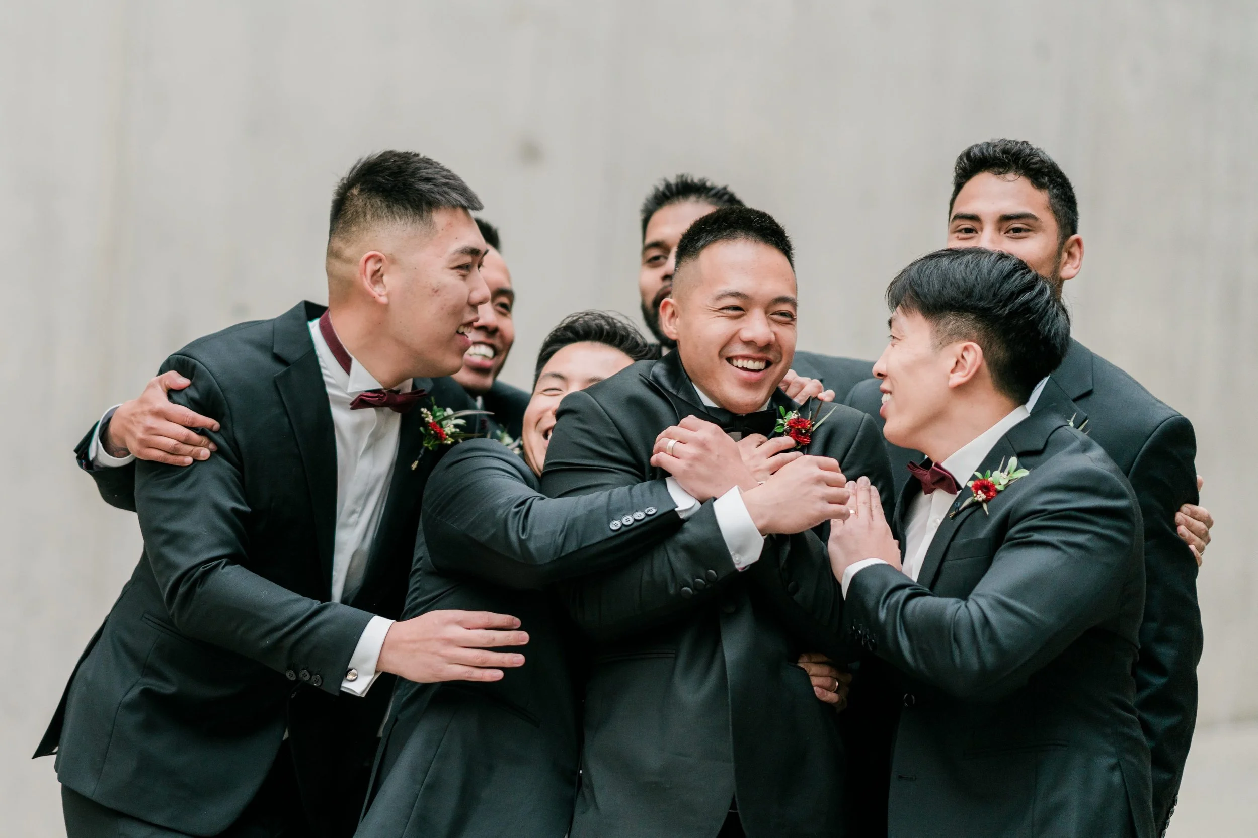 Group of men in tuxedos smiling and hugging each other at a wedding celebration.