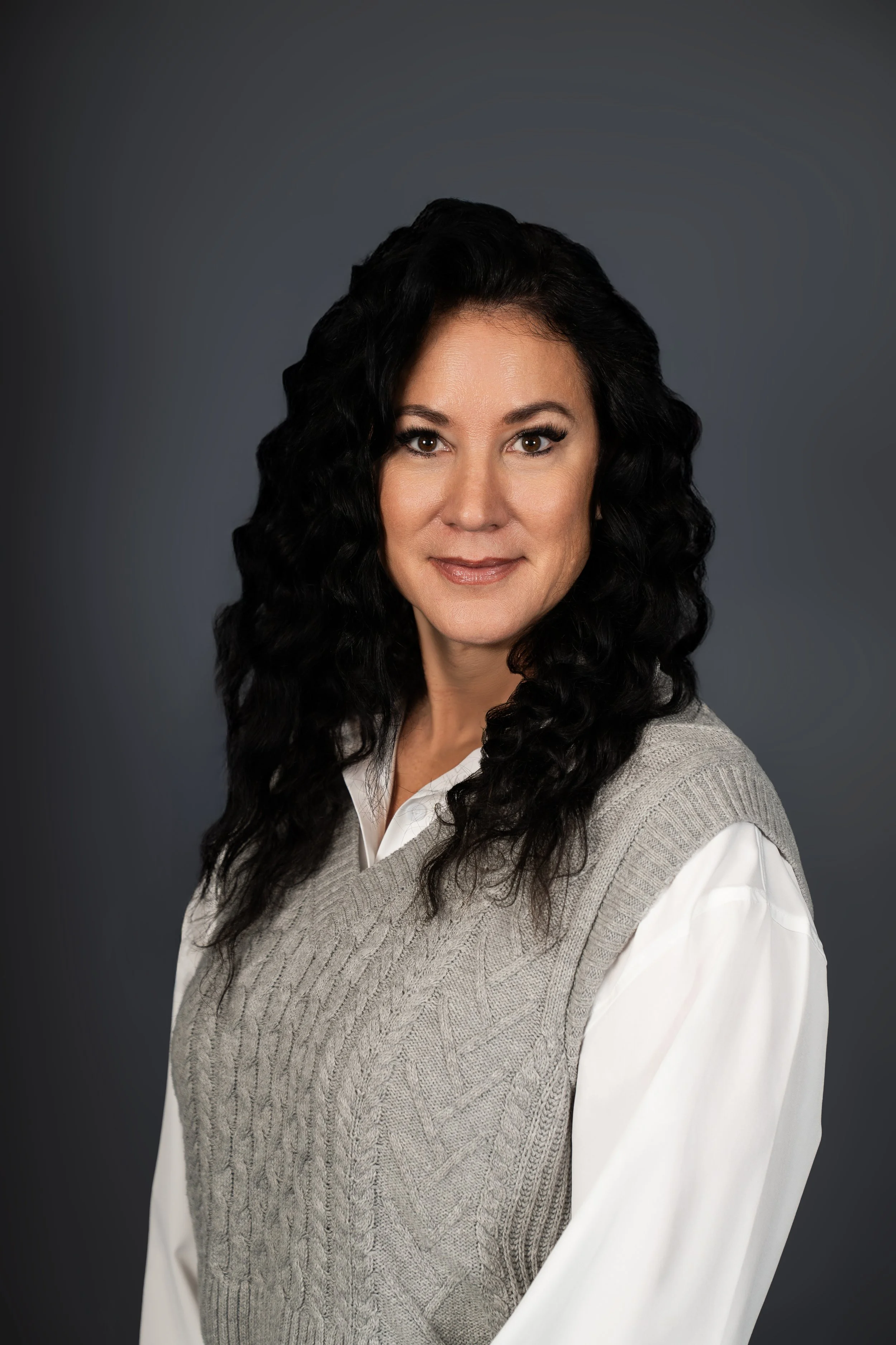 Portrait of a woman with curly black hair, wearing a white shirt and a gray knitted vest, against a dark gray background.