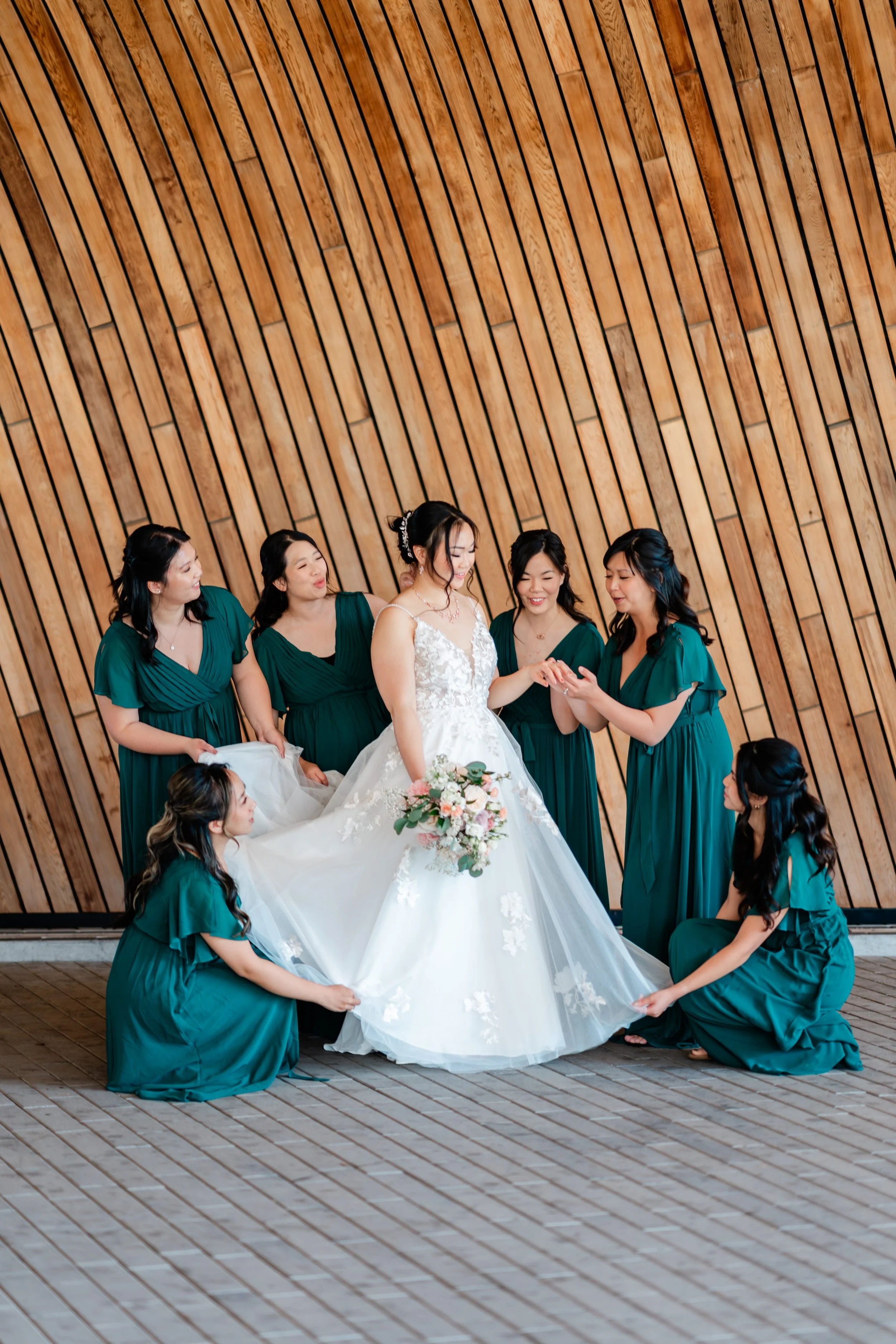 A bride in a white wedding gown holding a bouquet of flowers, surrounded by six women in dark green dresses, in front of a wooden wall.